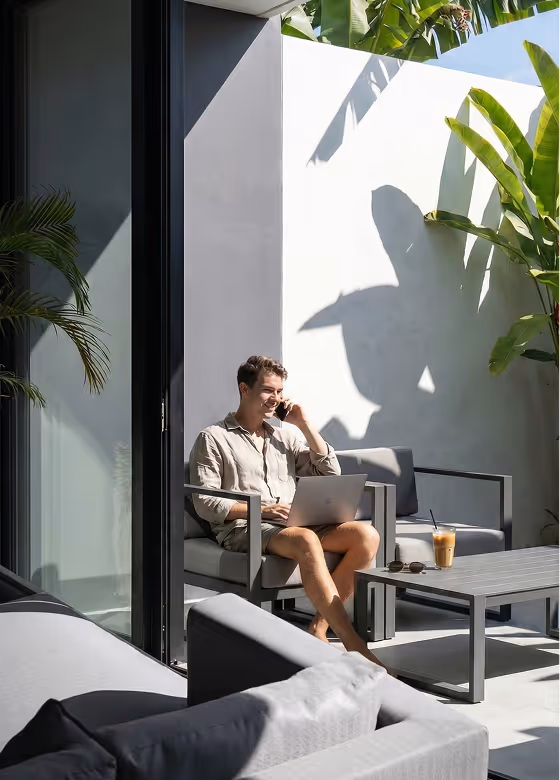A man sitting on a patio chair with a laptop on his lap, talking on a phone, surrounded by gray outdoor furniture and tropical plants.