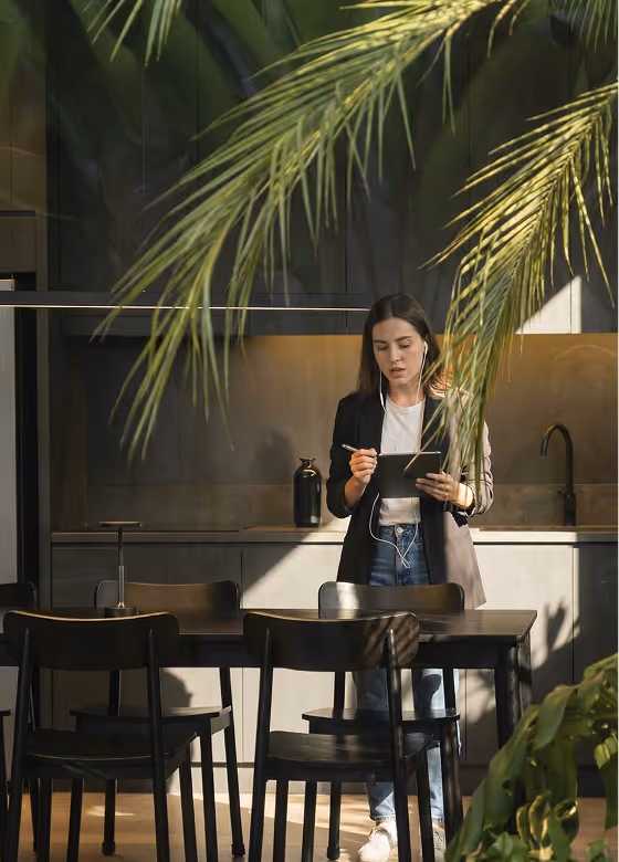 A woman in a black jacket with headphones stands at a table near the kitchen, fiddling with a tablet.