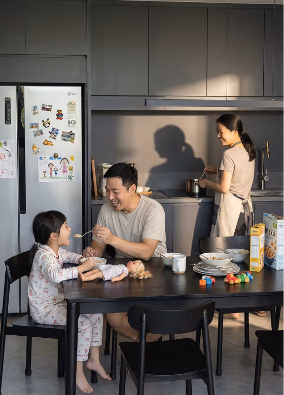 Father feeding daughter at kitchen table while mother cooks in background, with cereal boxes and toys on table.