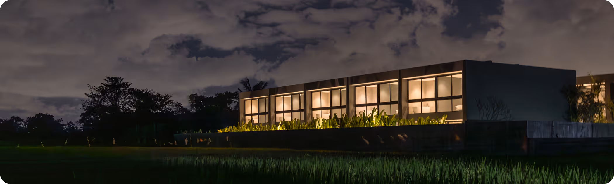 Modern building with large illuminated windows at night surrounded by greenery and trees under a cloudy sky.