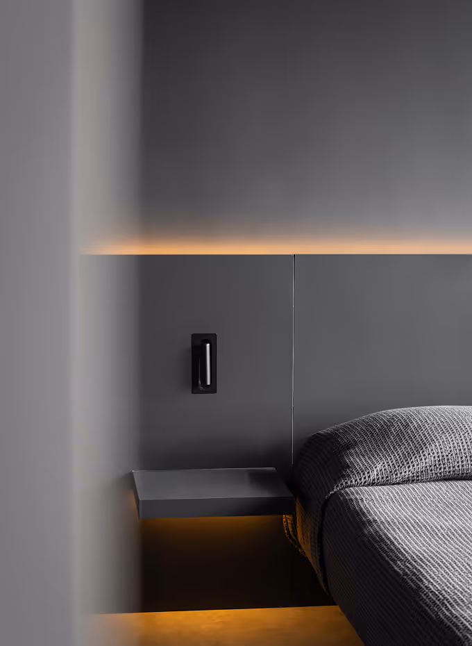 Minimalist bedroom corner with a floating bedside shelf, textured gray bedding, dark gray wall panel, and warm ambient lighting.
