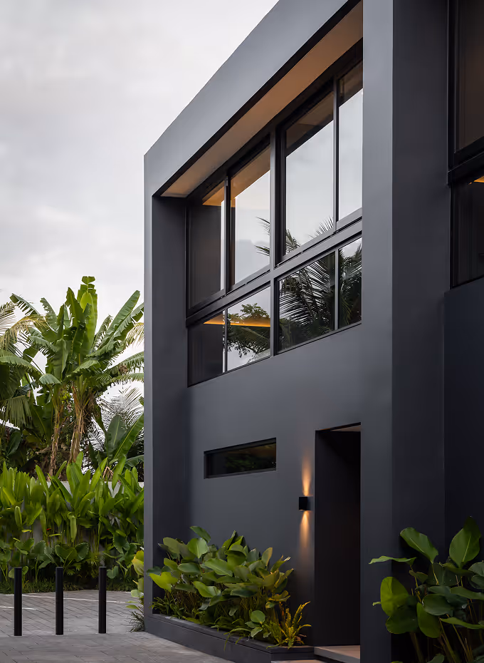 Modern grey building facade with large windows reflecting greenery and outdoor plants near the entrance.