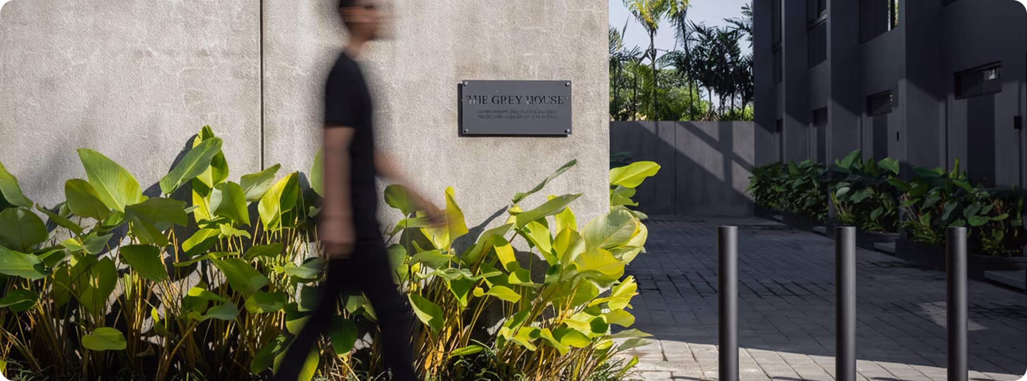 Blurred person walking past a concrete wall with a plaque that reads 'The Grey House' surrounded by green plants and modern building elements in the background.