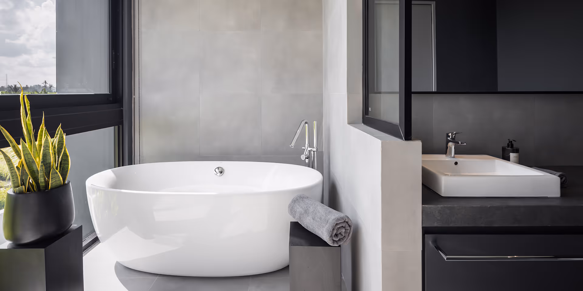 Modern bathroom with a white oval freestanding bathtub, grey towel on a black stool, and a white sink with a black countertop.