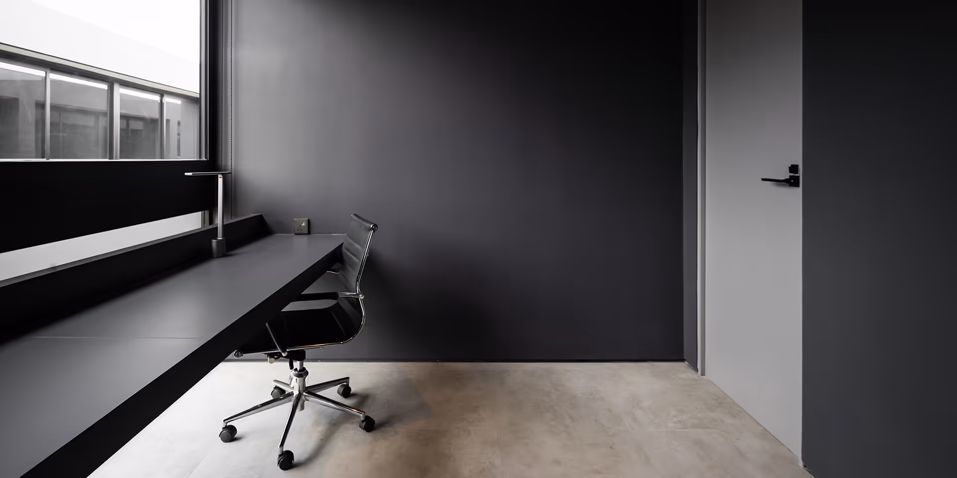 Minimalist office corner with a black desk, black swivel chair, gray wall, and window letting in natural light.