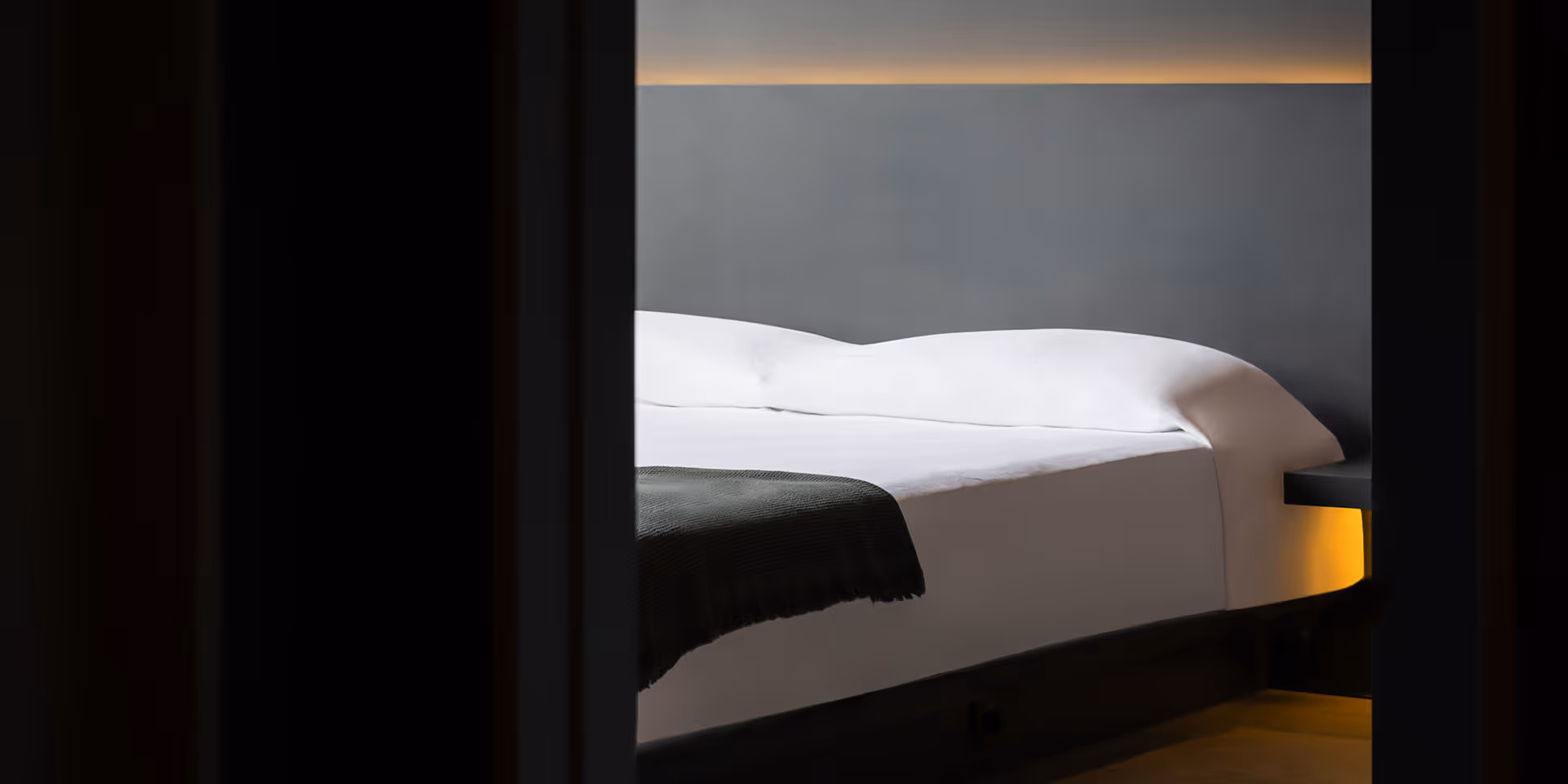 Minimalist bedroom with a white bed, dark gray headboard wall, and warm ambient lighting.