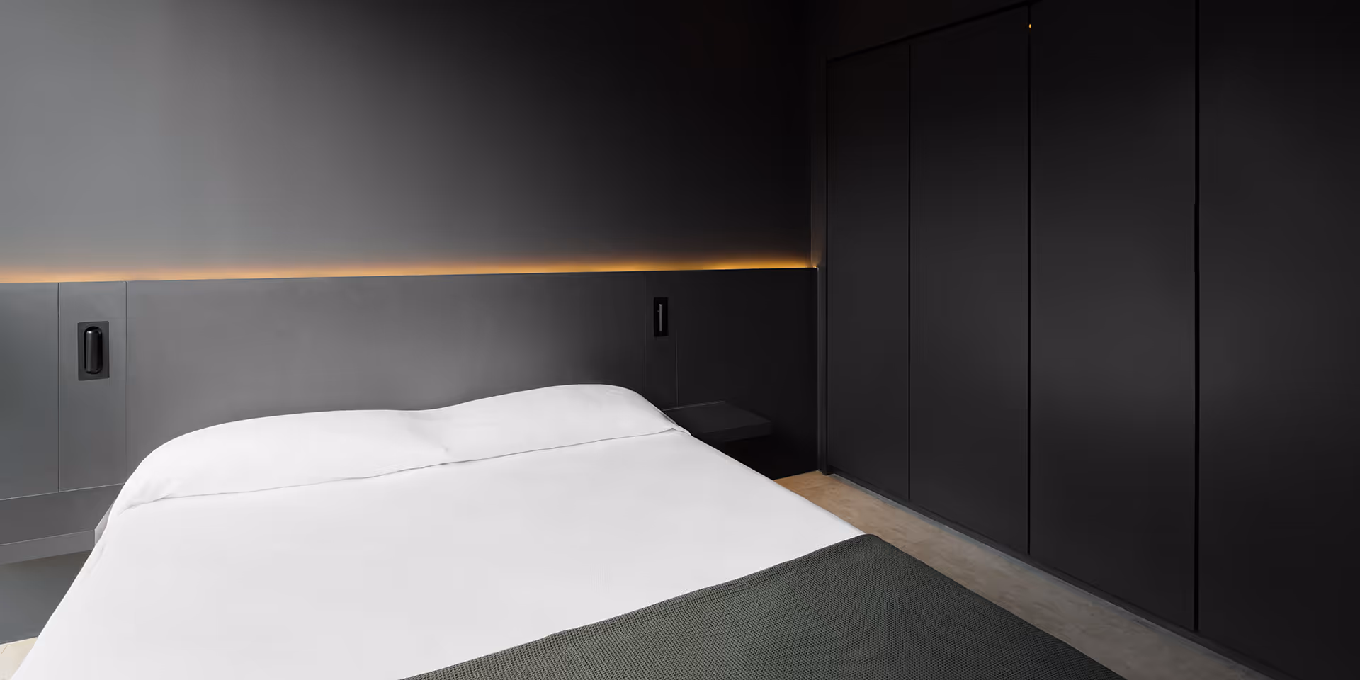Minimalist bedroom with a bed featuring white sheets, dark gray blanket, dark gray wall panels, and soft orange lighting above the headboard.