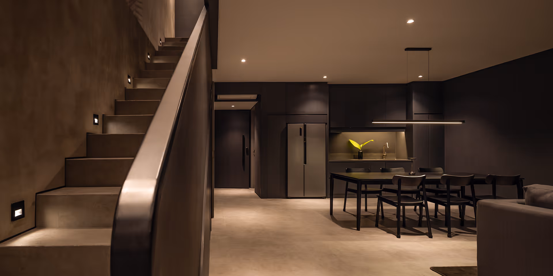 Modern dark-toned kitchen and dining area with black chairs, a table, a refrigerator, and illuminated stairs on the left.