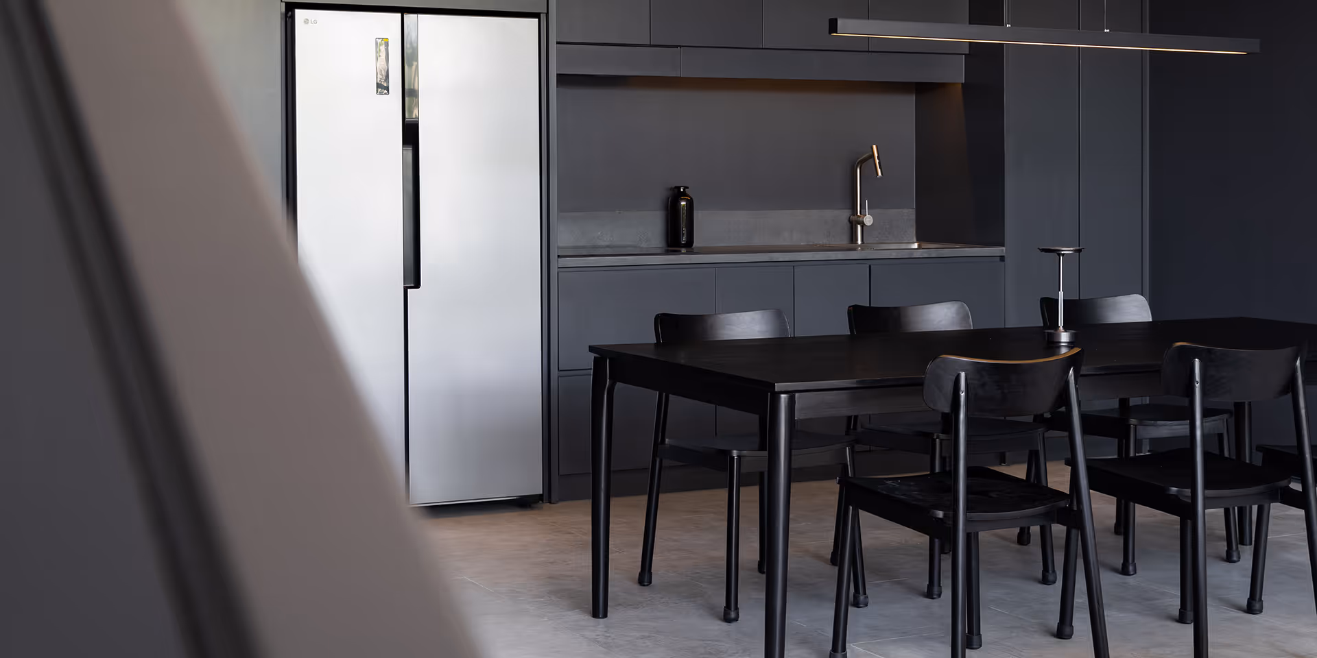 Minimalist kitchen with matte black cabinets, a silver LG refrigerator, black dining table, and six black chairs under a long linear pendant light.