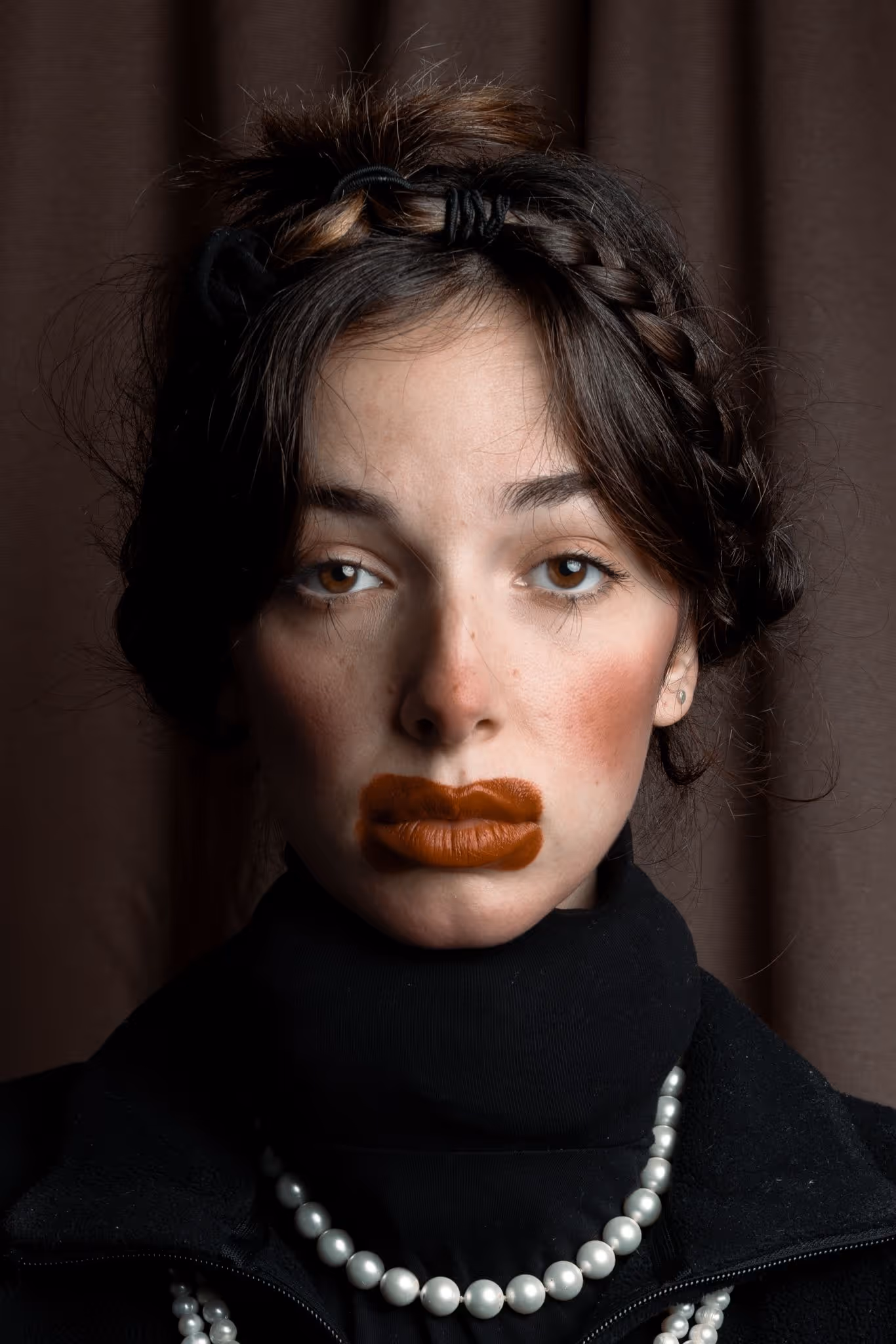 Mujer con labios pintados de color marrón fuera del contorno natural, cabello trenzado y collar de perlas.