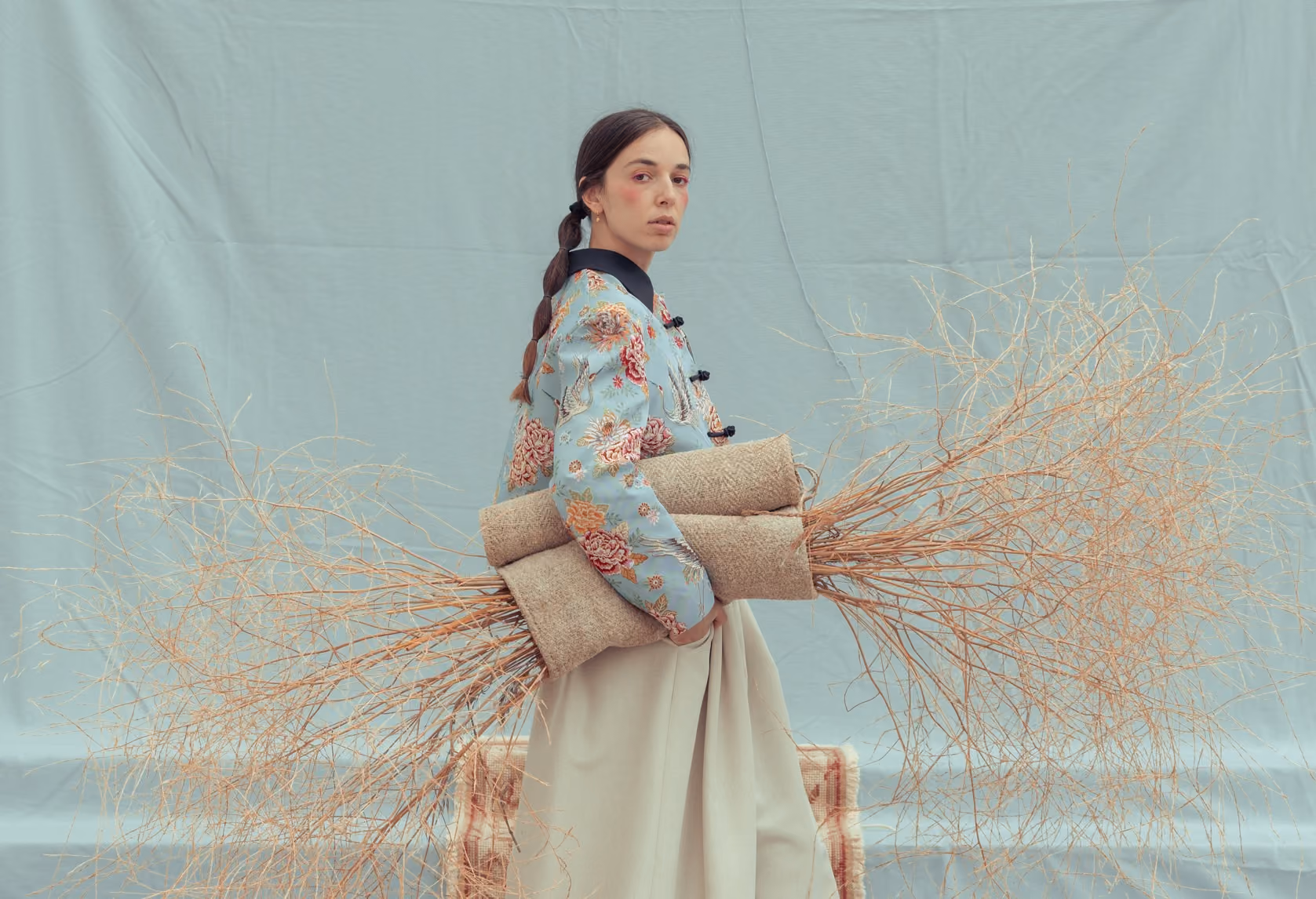 Mujer con chaqueta florar y falda beige sostiene ramas secas enrolladas en alfombras.