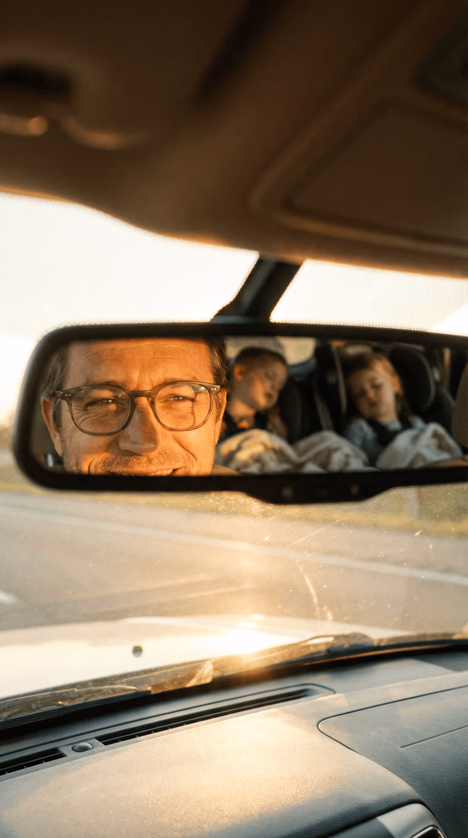 Smiling man with glasses reflected in car rearview mirror with two children sleeping in car seats behind him.