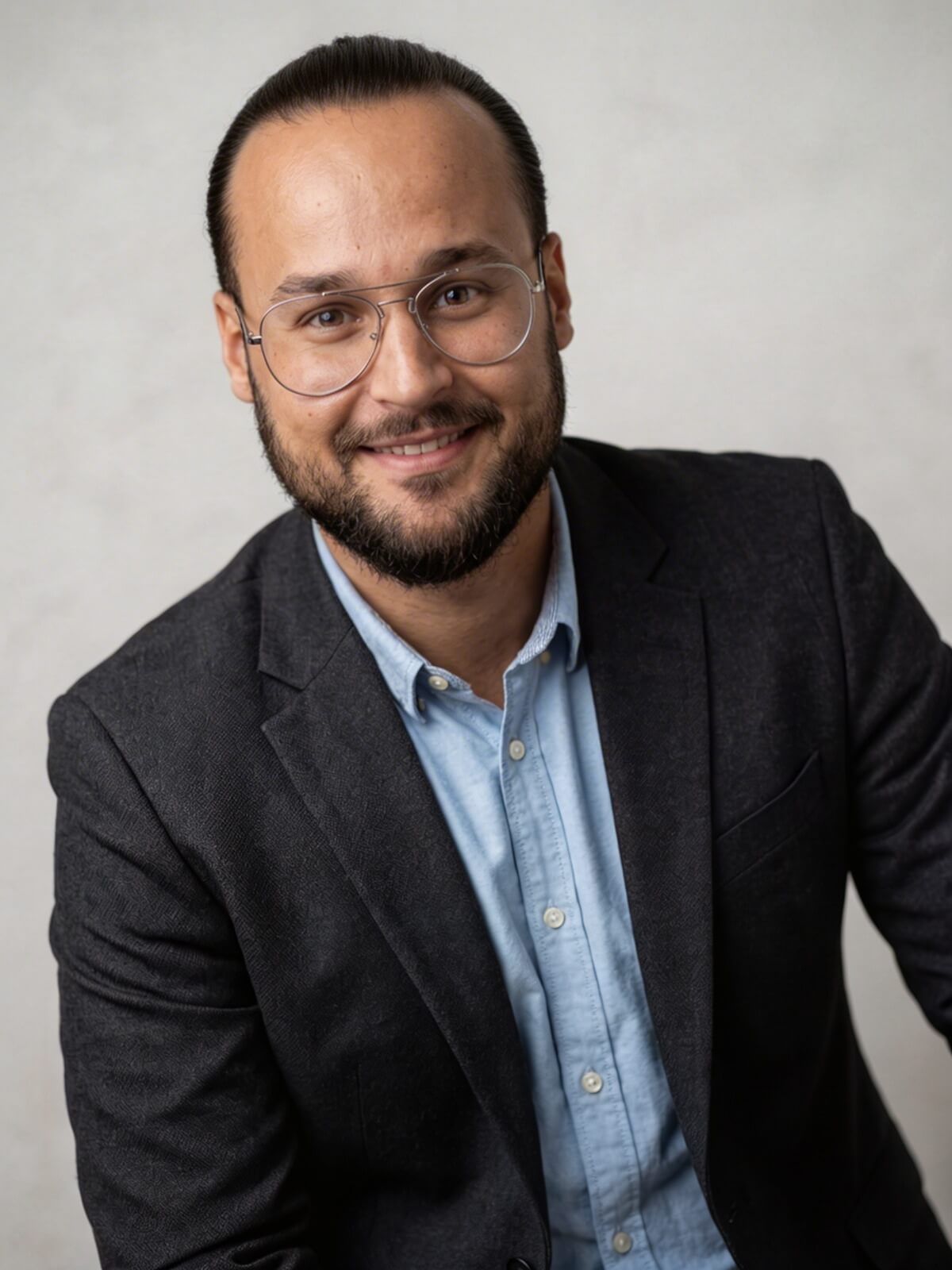 Smiling man with beard and glasses wearing a dark blazer over a light blue shirt.