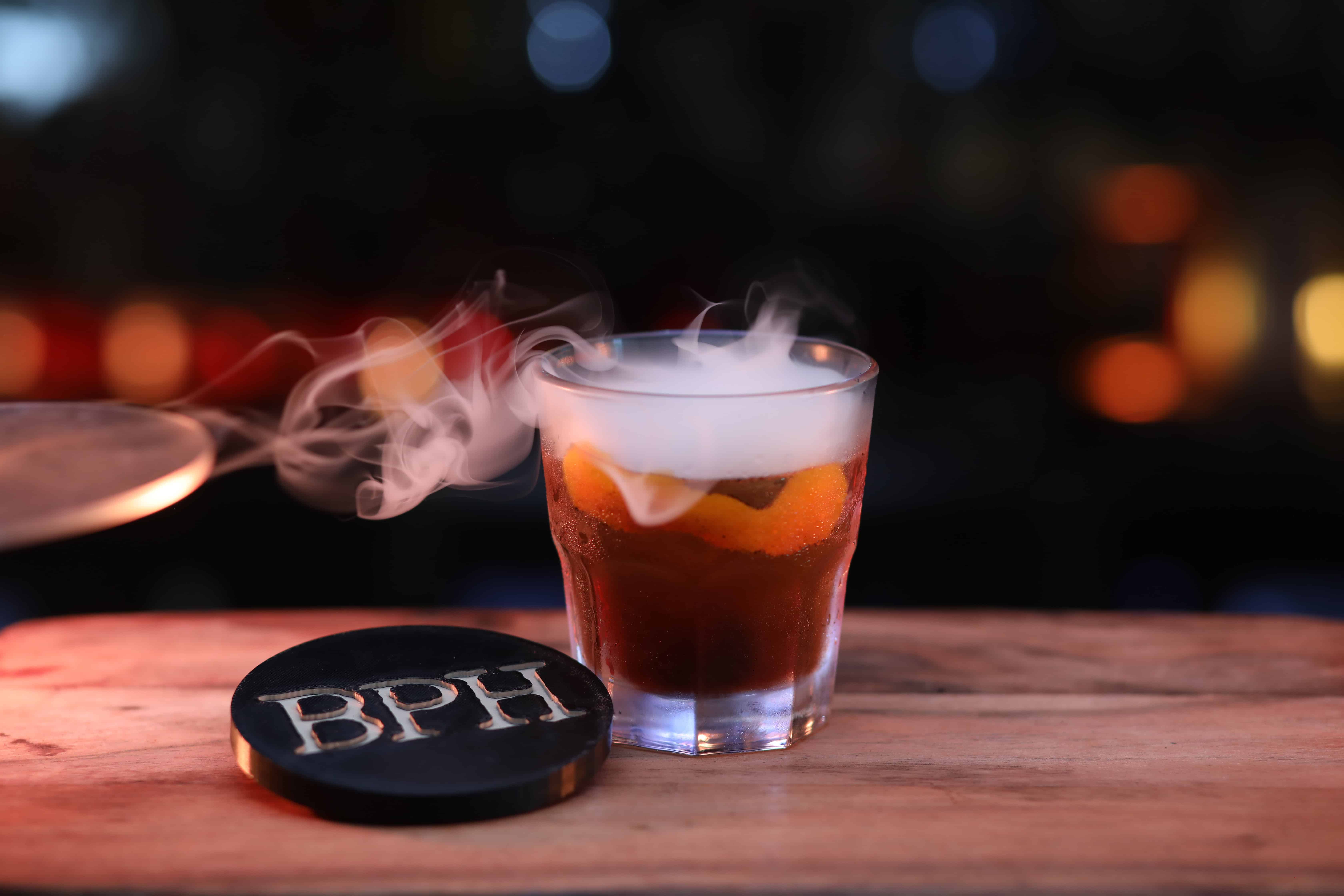 Classic Old Fashioned cocktail in a rocks glass with a large clear ice cube and orange peel twist. Dark moody bar setting.