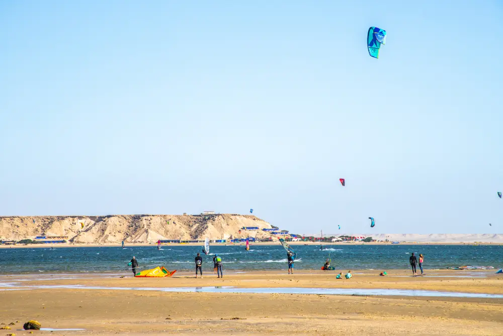Several kitesurfers and kites over the flat Dakhla lagoon on a sunny, windy afternoon with the desert plateau behind