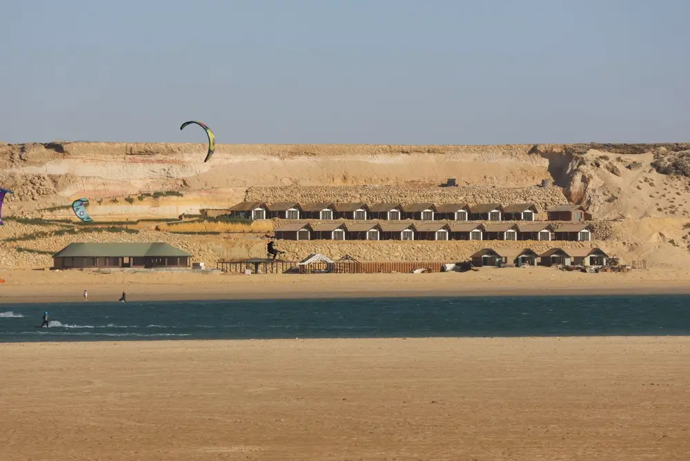 Rows of white-shuttered bungalows at a Dakhla lagoon camp below sand cliffs, with two kitesurfers riding the lagoon in front
