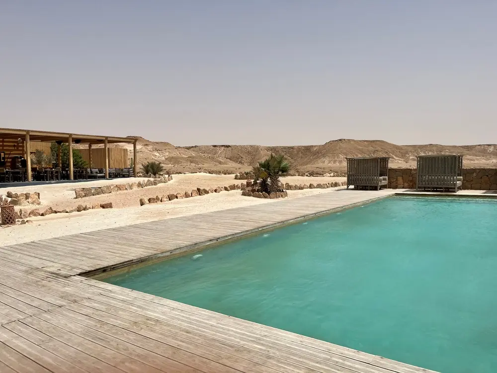 Turquoise pool and wooden decking at a Dakhla desert camp, with a shaded lounge area and rocky ridges behind
