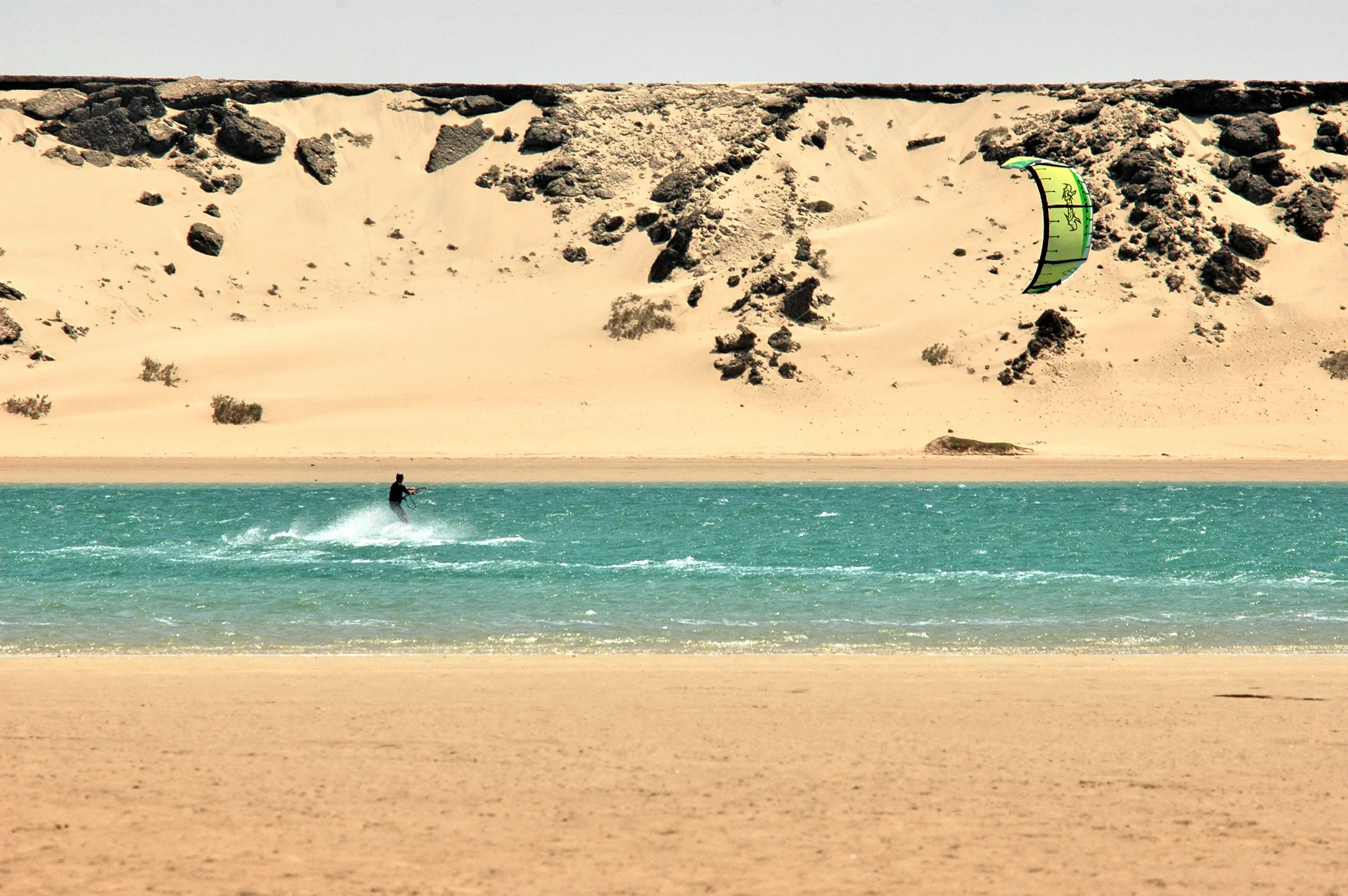 Kitesurfer passing below the White Dune on the Dakhla lagoon, pale sand wall rising above flat turquoise water