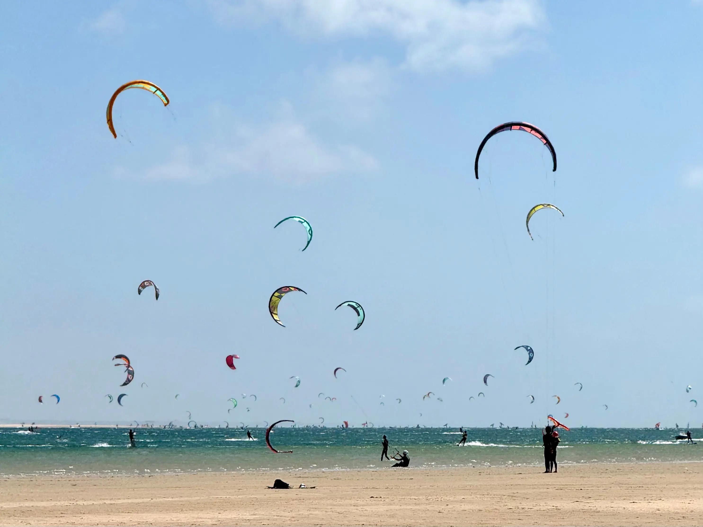 Dozens of kites in the sky above the Dakhla lagoon on a busy peak-season day, riders on the water beyond the beach