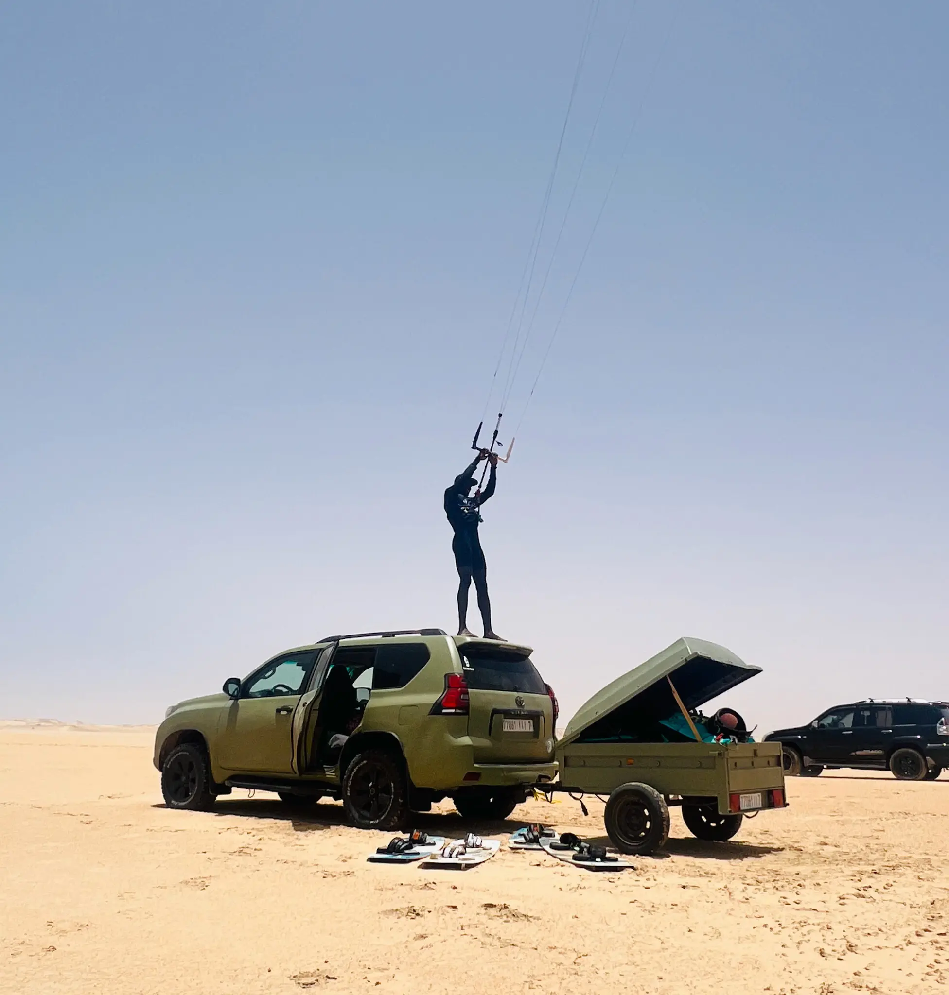 Kitesurf guide standing on a 4x4 roof holding a kite bar, gear and trailer laid out on the sand before a Dakhla downwinder