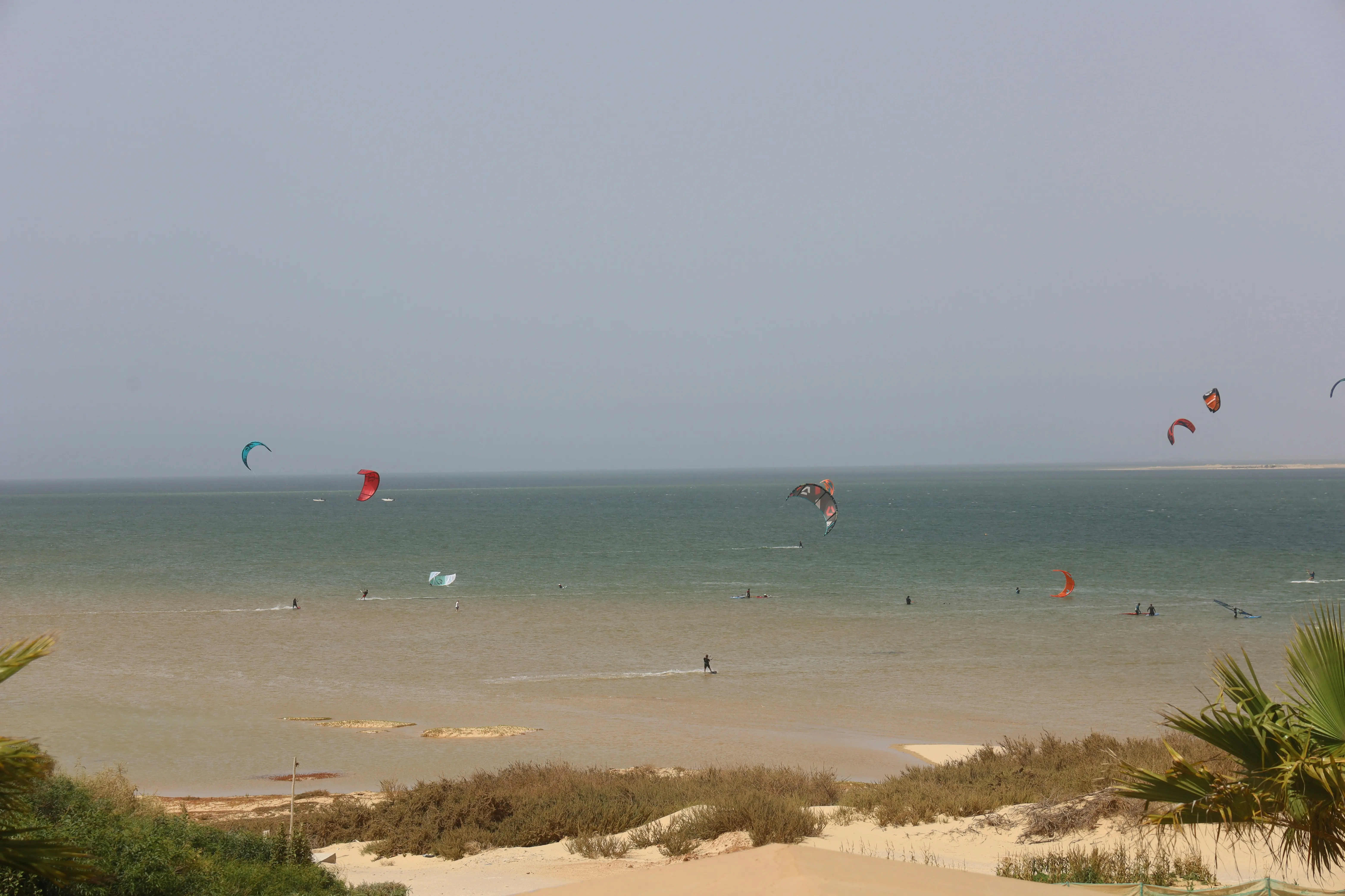 Wide view across the Dakhla lagoon from the inner shore, kites spread over shallow water with a sandbar on the far side