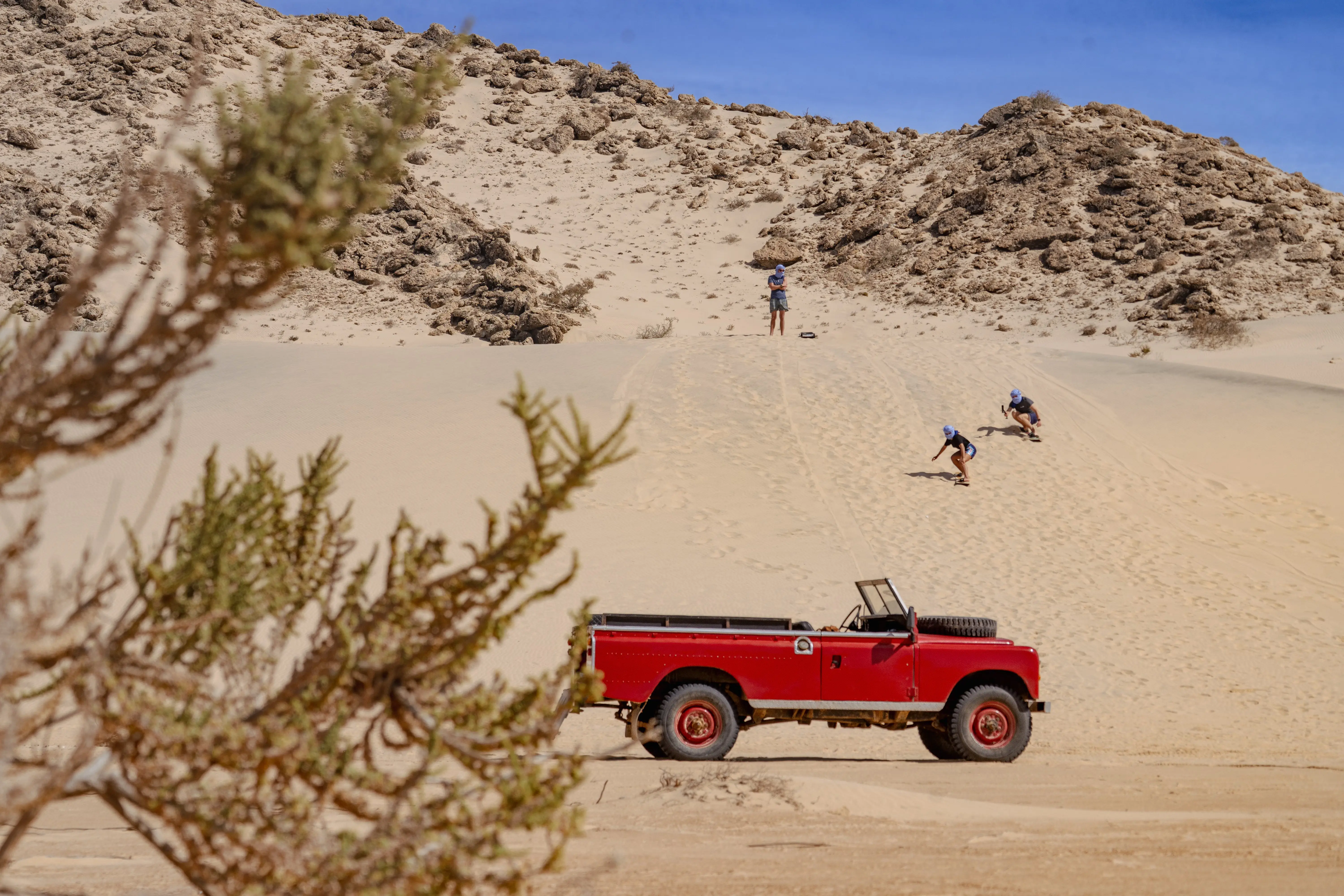 Three people sandboarding down a dune in the Dakhla desert with a vintage red Land Rover parked at the foot of the slope