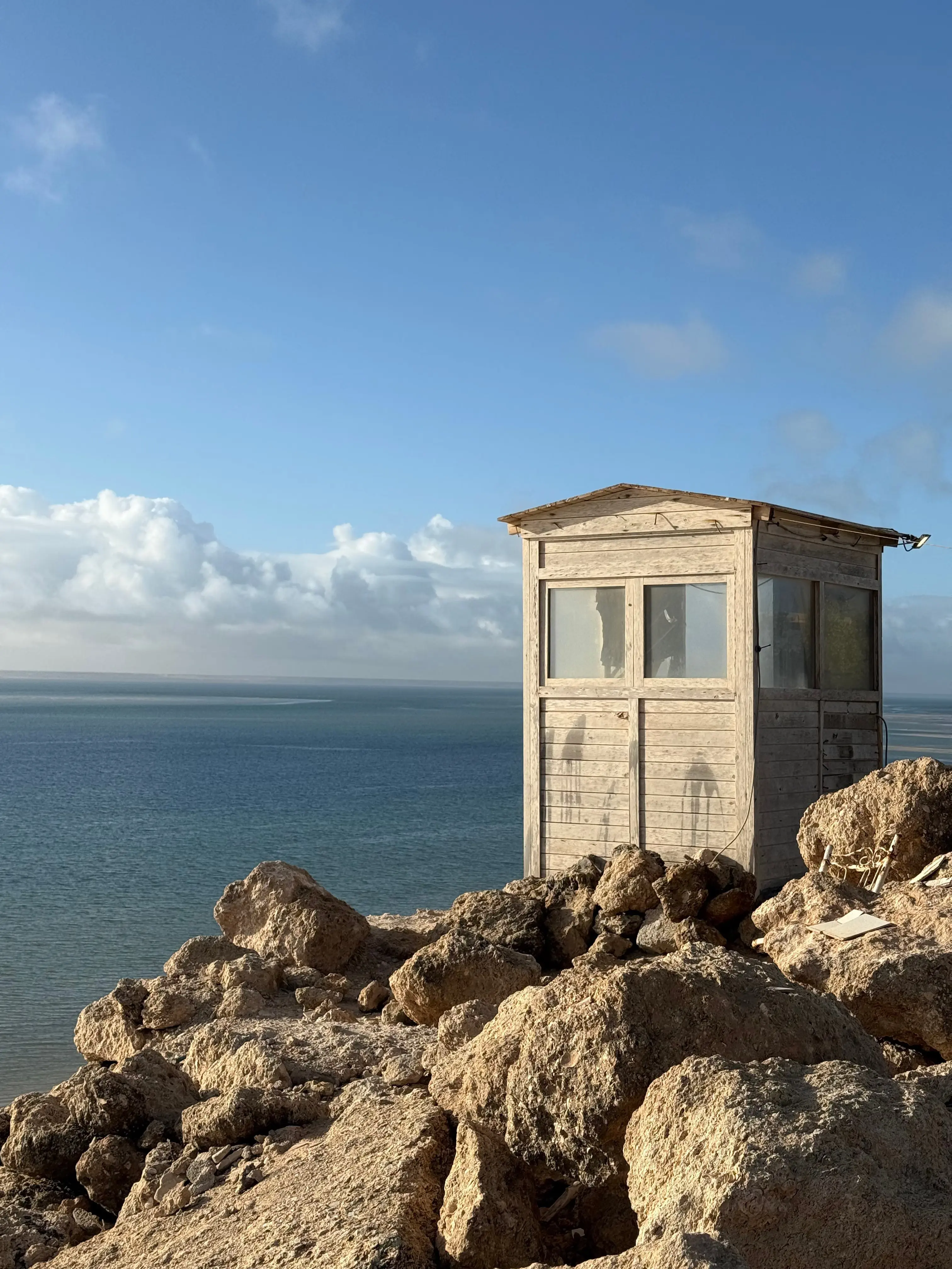 A weathered wooden hut on the rocky Atlantic shoreline near Dakhla, with flat calm water beyond