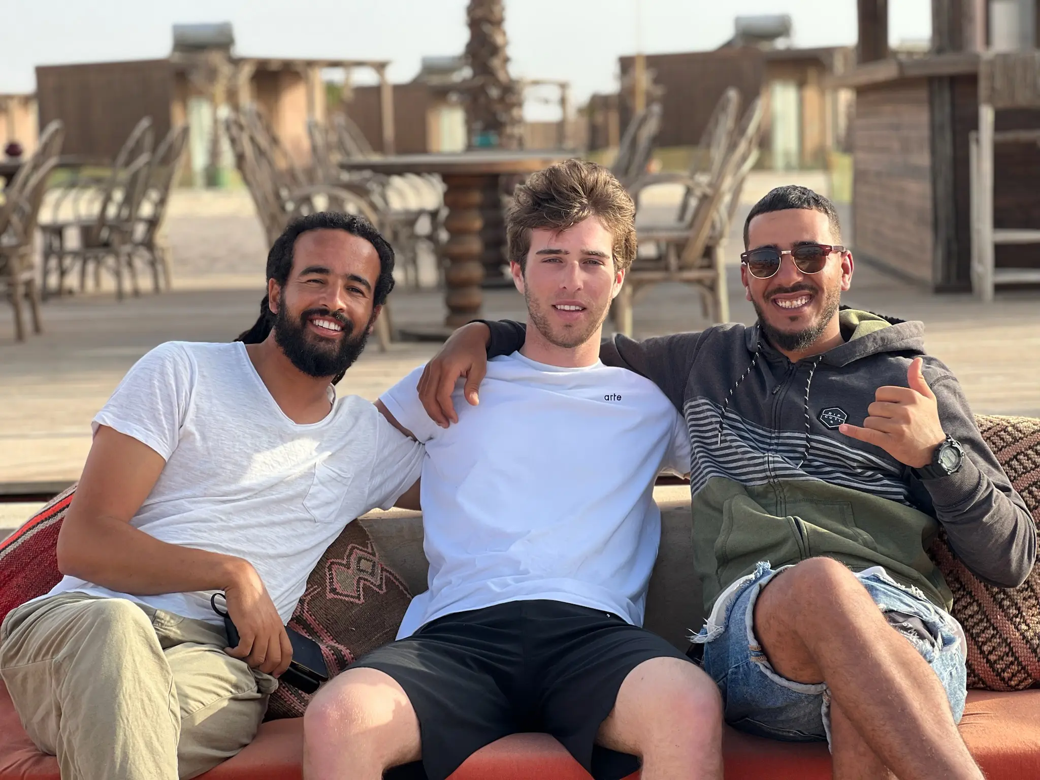 Three people sitting on a low sofa on a Dakhla camp terrace in late afternoon light, with camp furniture and a palm behind them