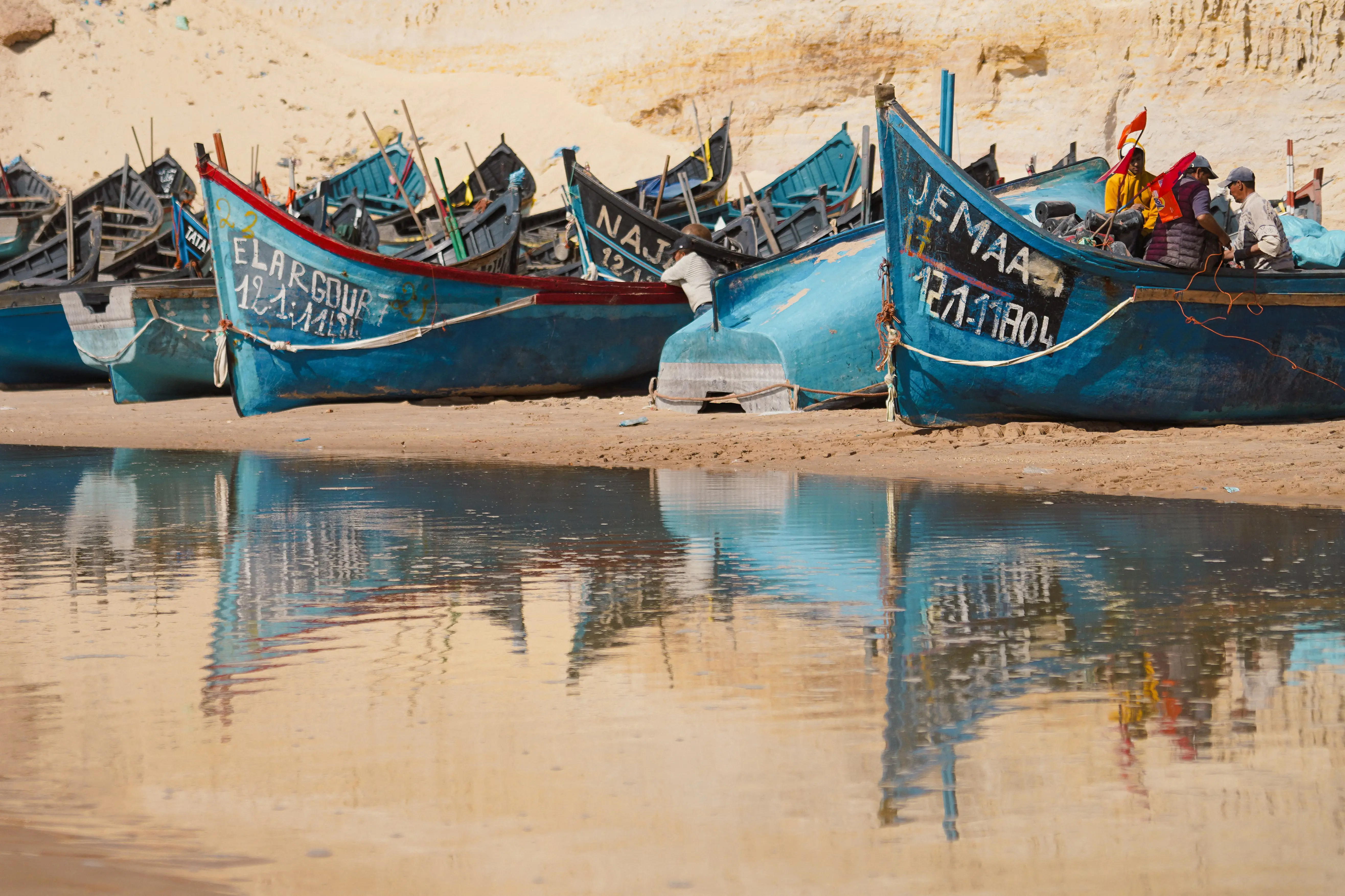 Blue wooden fishing boats drawn up on the sand at Dakhla, reflected in still shallow water, with fishermen working on one of them
