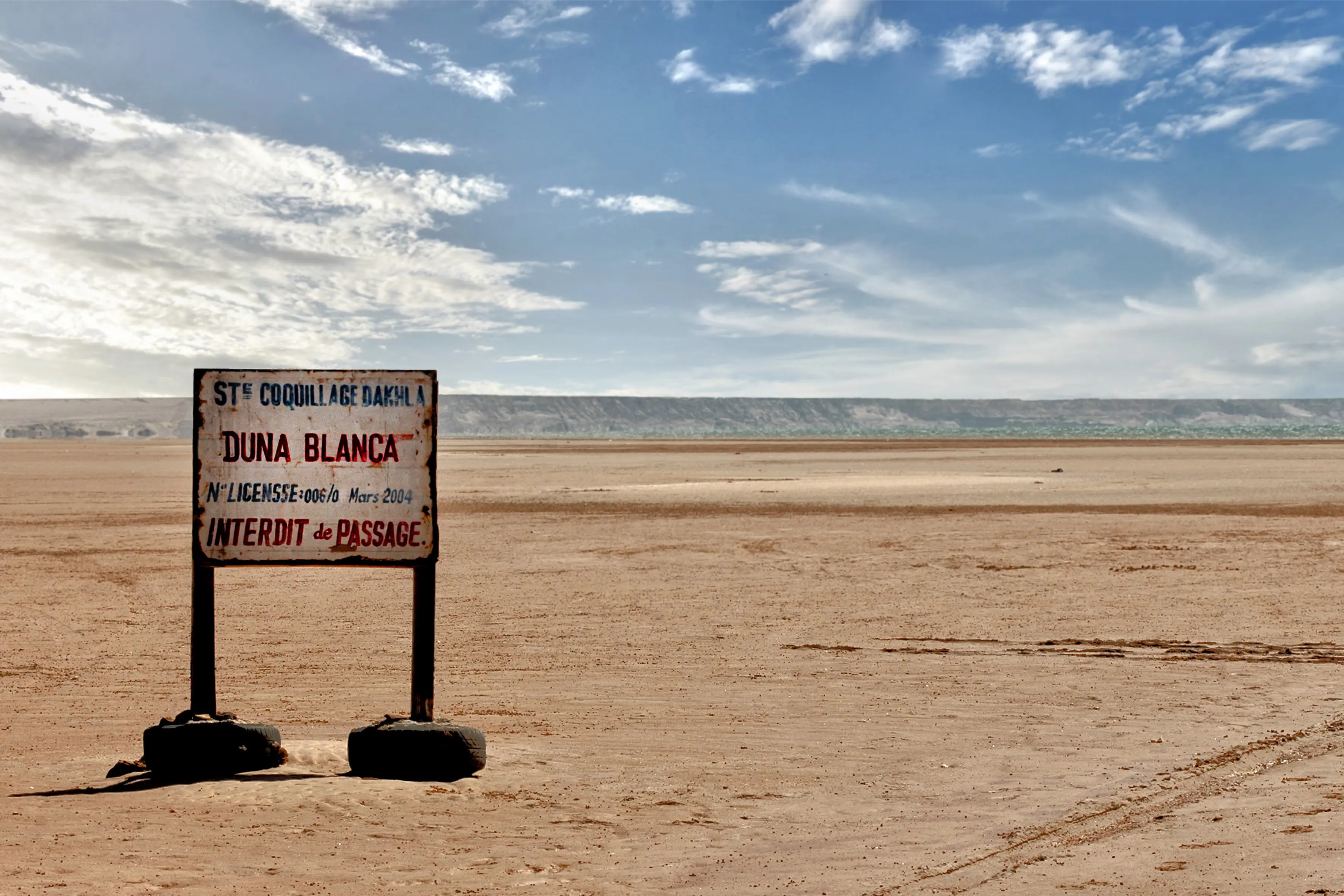 Weathered Duna Blanca sign standing in flat sand near Dakhla's White Dune, with the peninsula cliffs and the lagoon behind