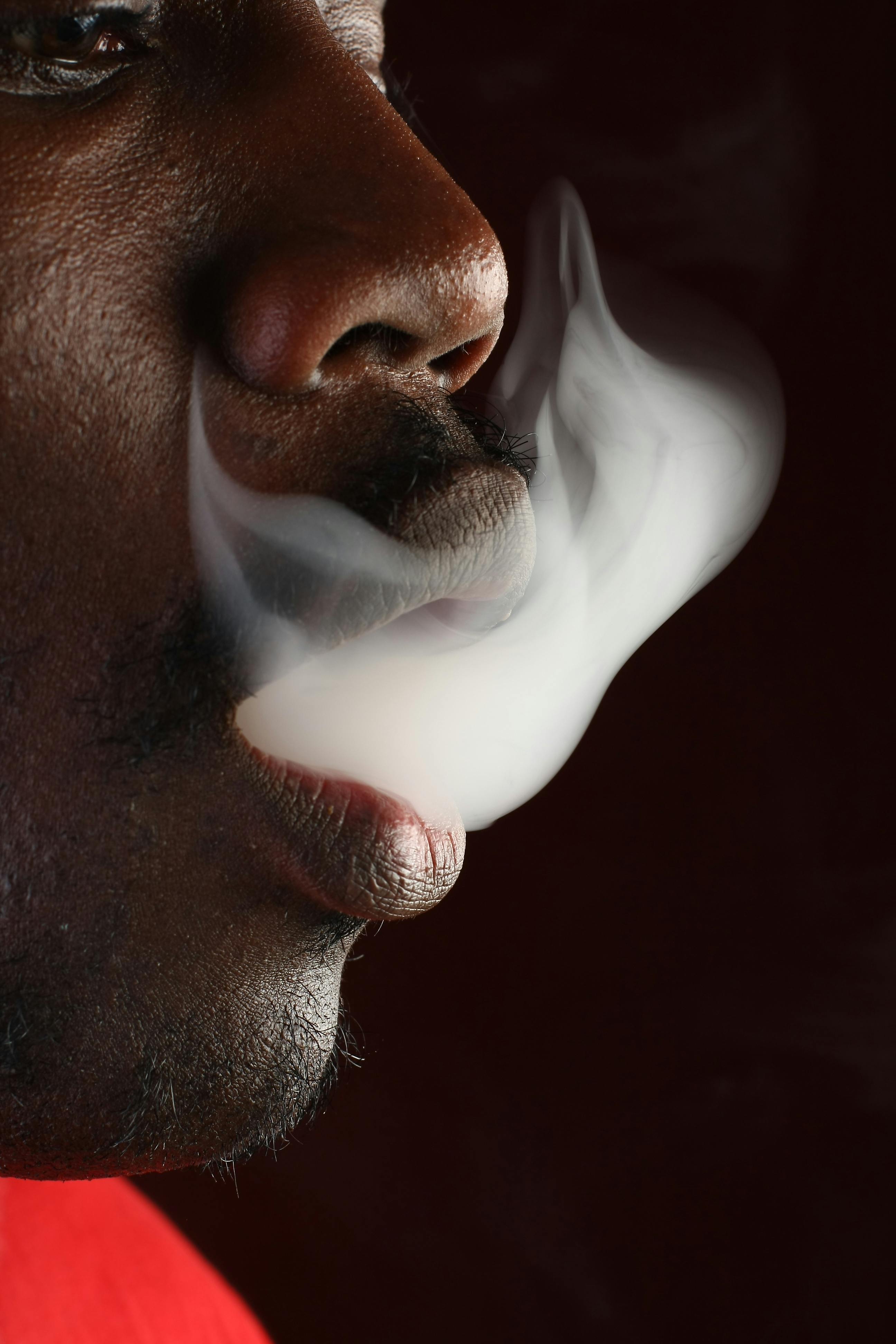 Close-up side view of a man exhaling white smoke against a dark background.