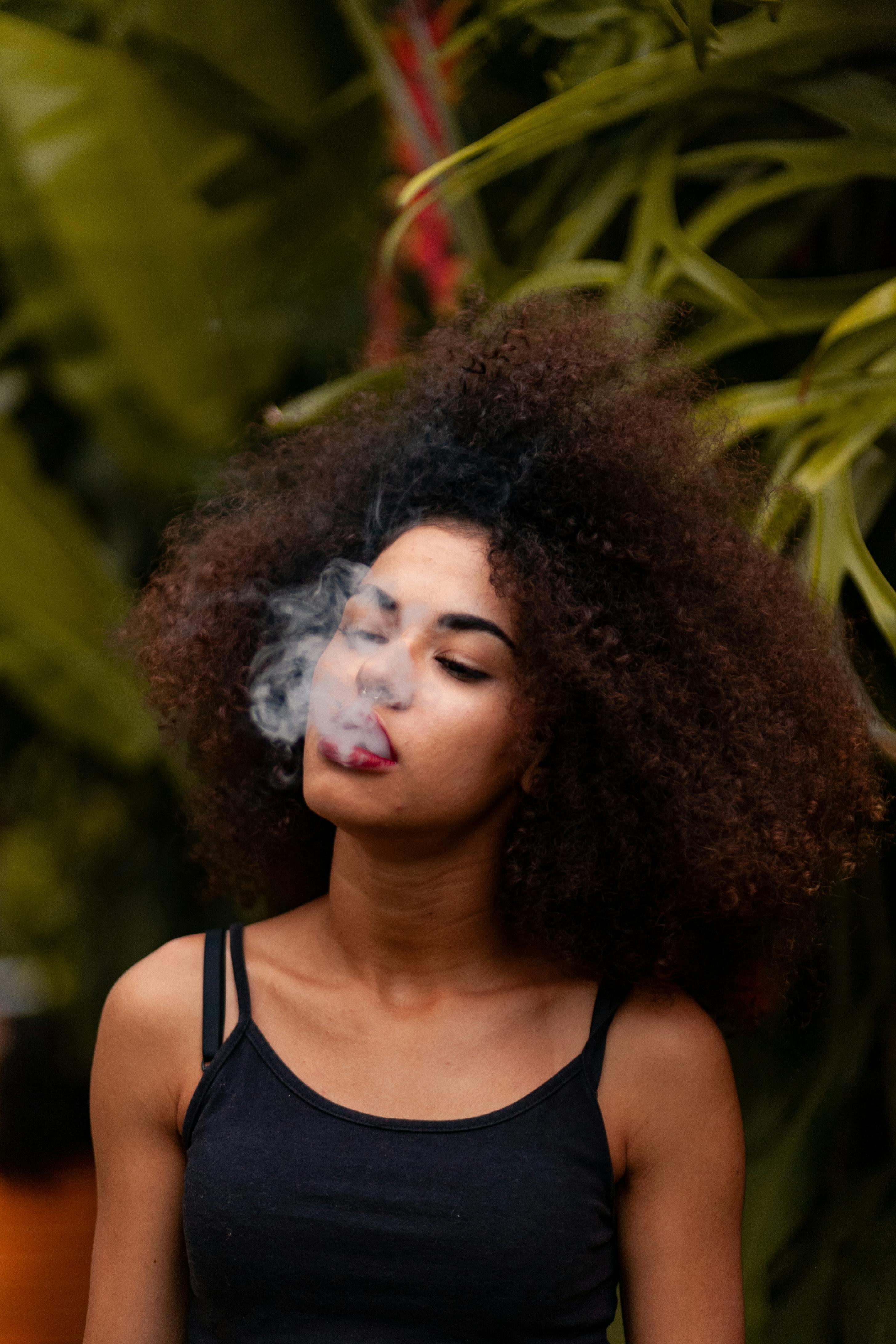 Young woman with voluminous curly hair wearing a black tank top, exhaling smoke outdoors with green foliage in the background.