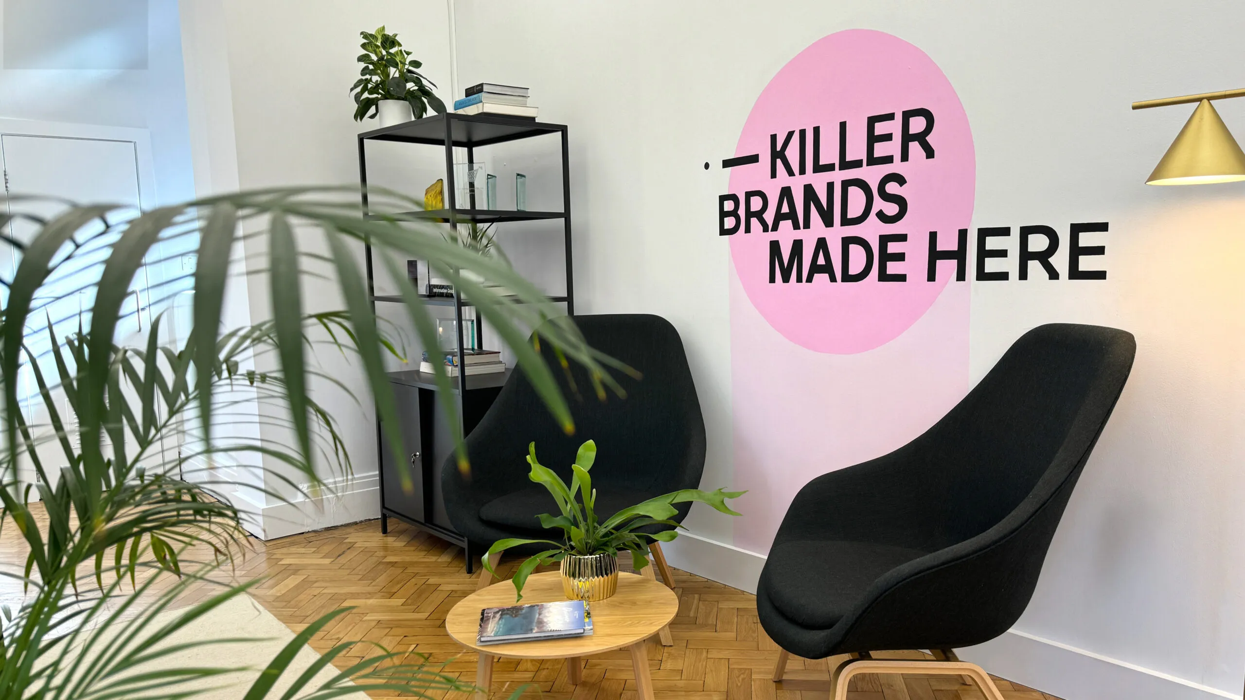 Modern office corner with two black chairs, wooden floor, green plants, and wall text 'KILLER BRANDS MADE HERE'.