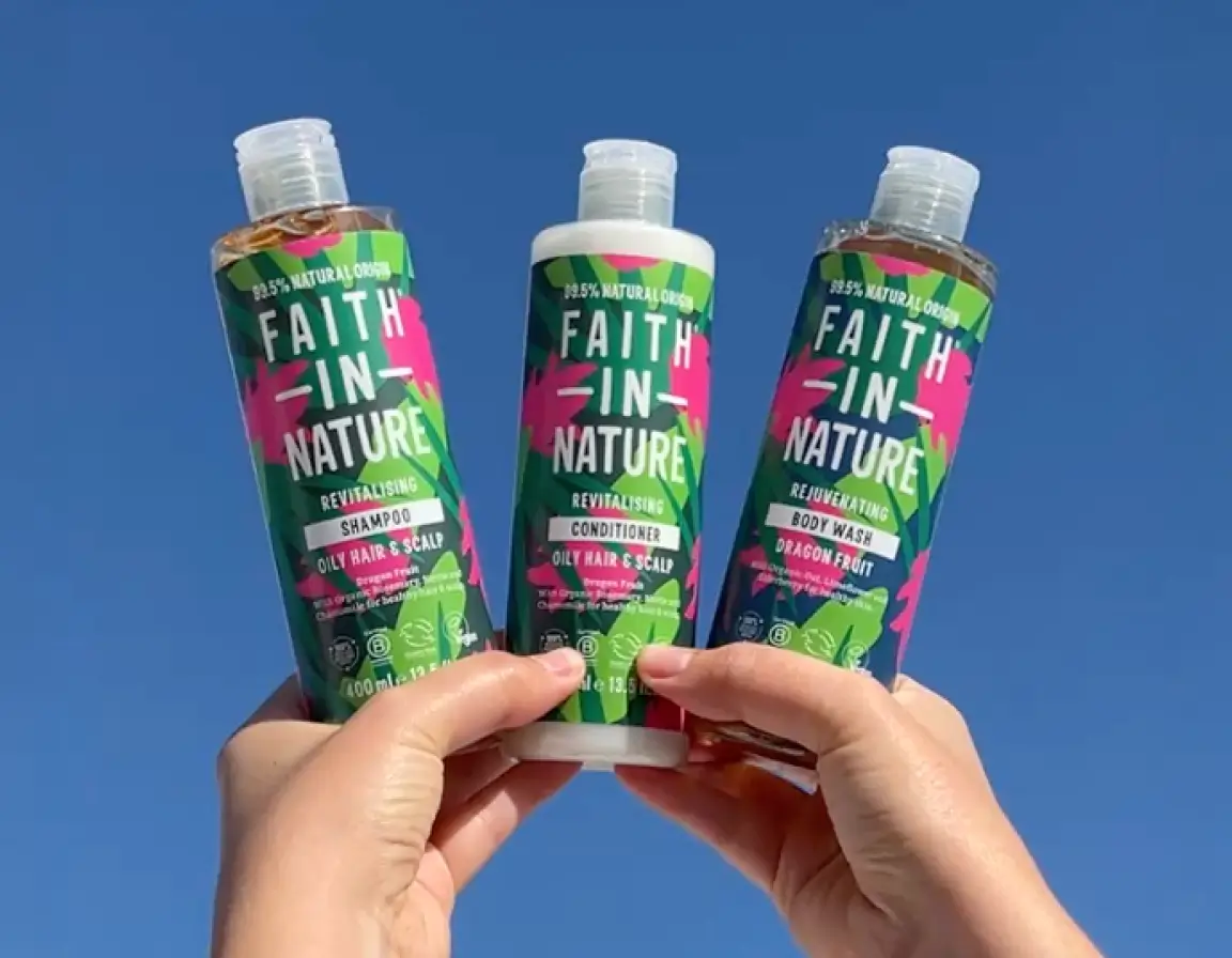 Hands holding three Faith In Nature products against a clear blue sky: revitalising shampoo, conditioner for oily hair and scalp, and dragon fruit body wash.