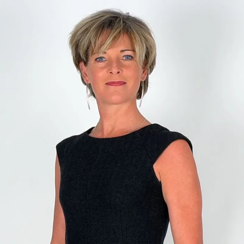 Confident woman with short blond hair wearing a sleeveless black dress against a plain background.