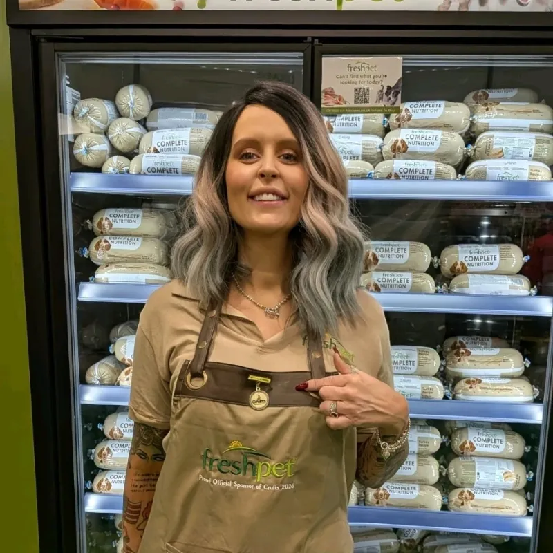 Woman with wavy hair wearing a Freshpet apron points to a Crufts 2026 badge in front of a refrigerator stocked with Freshpet Complete Nutrition dog food.
