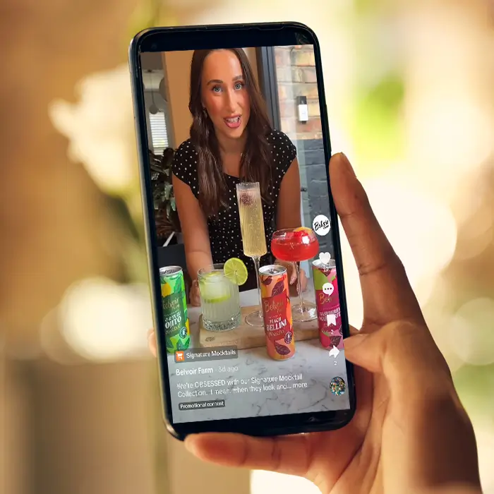 Hand holding smartphone showing a woman presenting canned and glass mocktails on a video.