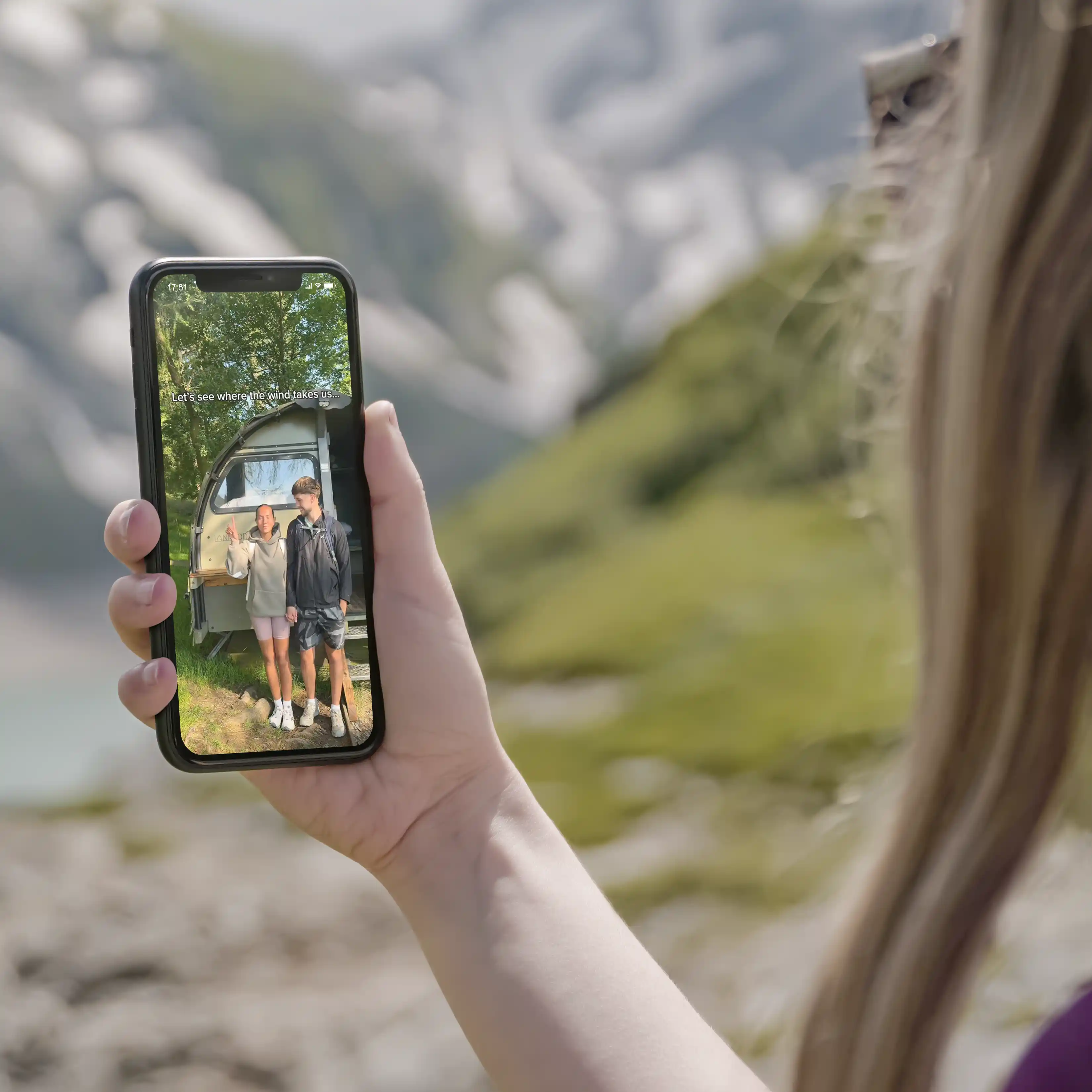 Person holding phone showing video of a couple standing outdoors near a caravan in a green mountainous area.