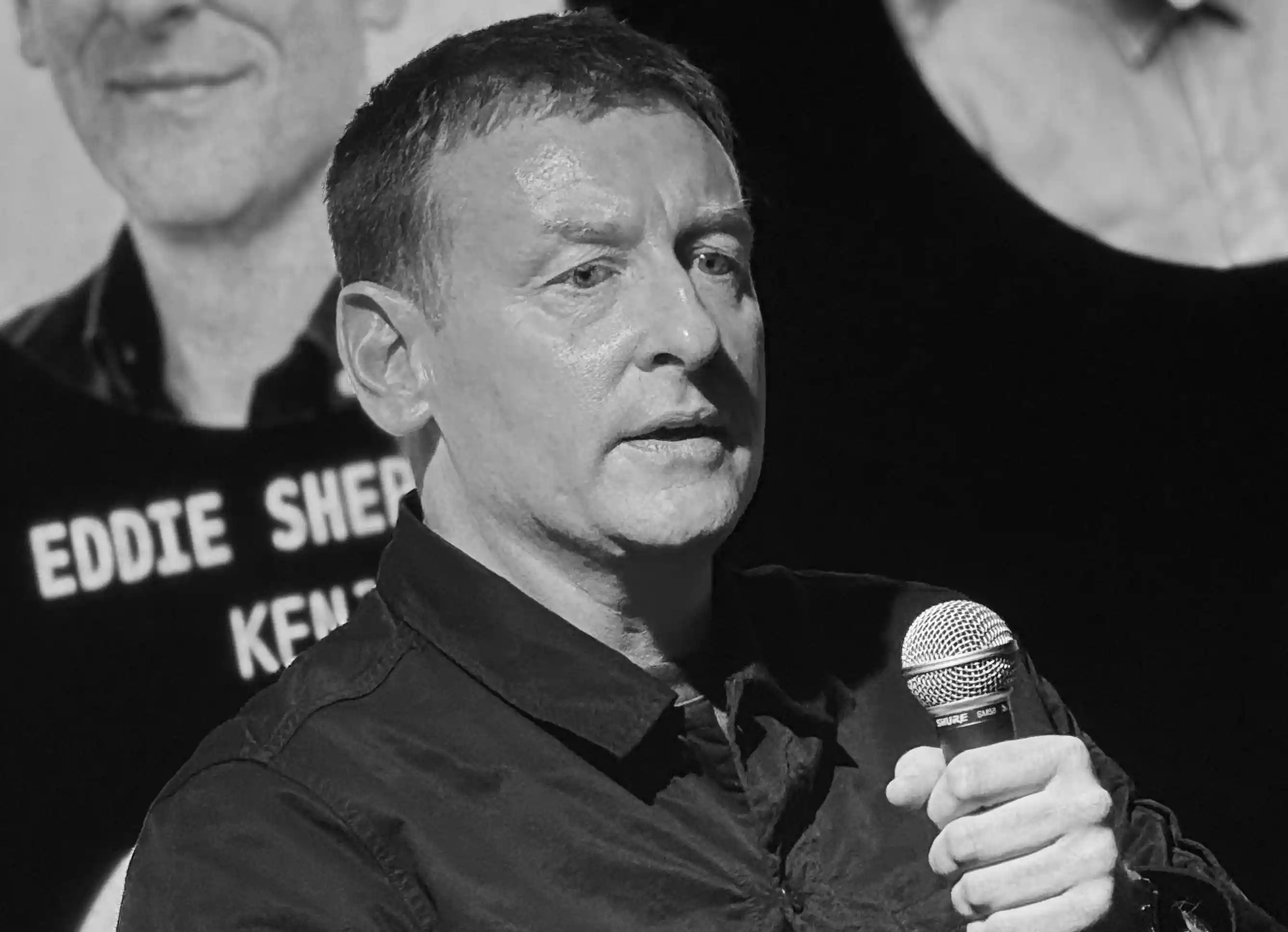Man speaking into a microphone in front of a screen showing the name Eddie Shepard Kent.