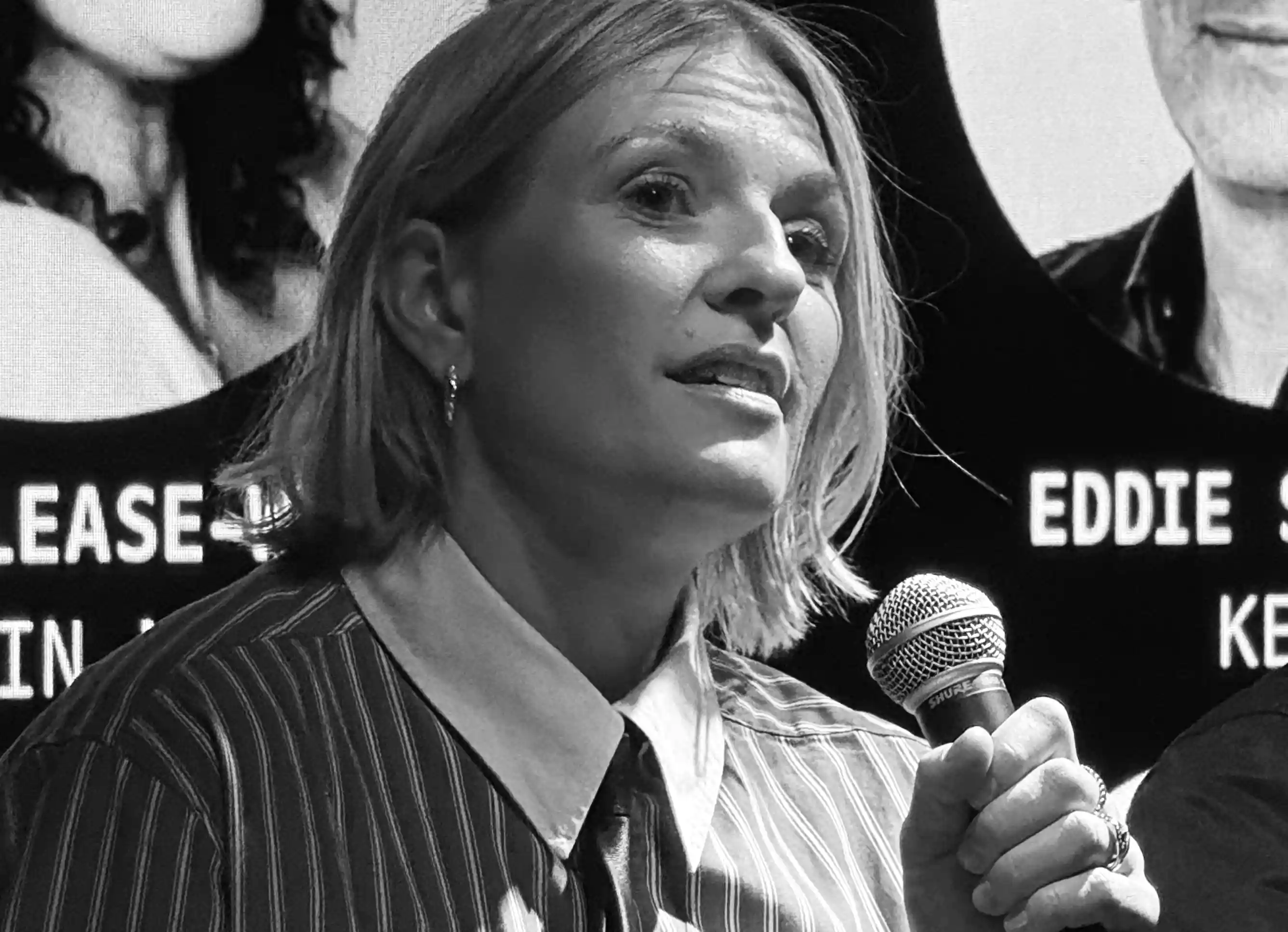 Woman holding microphone speaking, wearing striped shirt and tie against a backdrop with partially visible text.