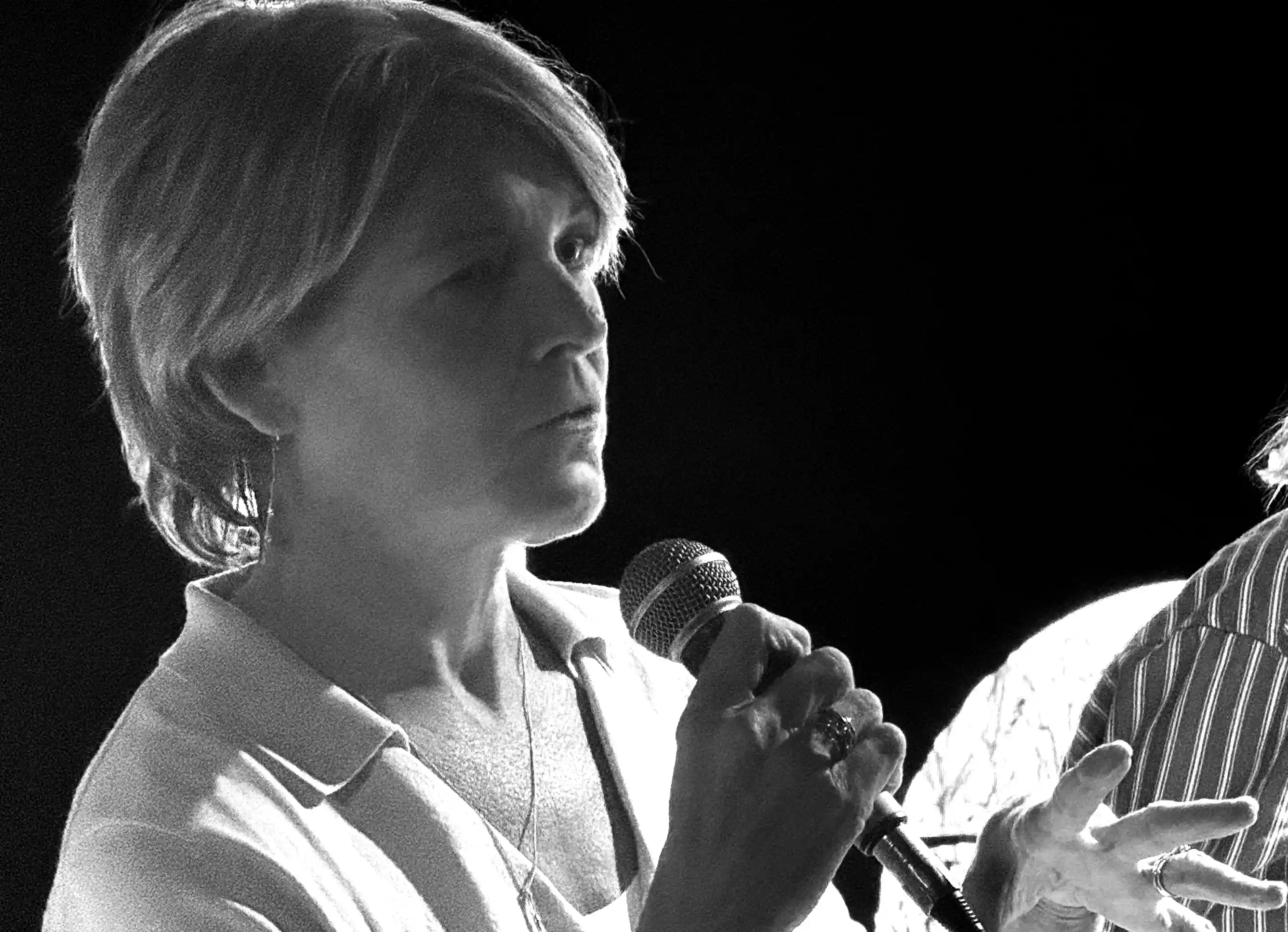 Woman holding a microphone speaking with expressive hand gestures against a dark background.