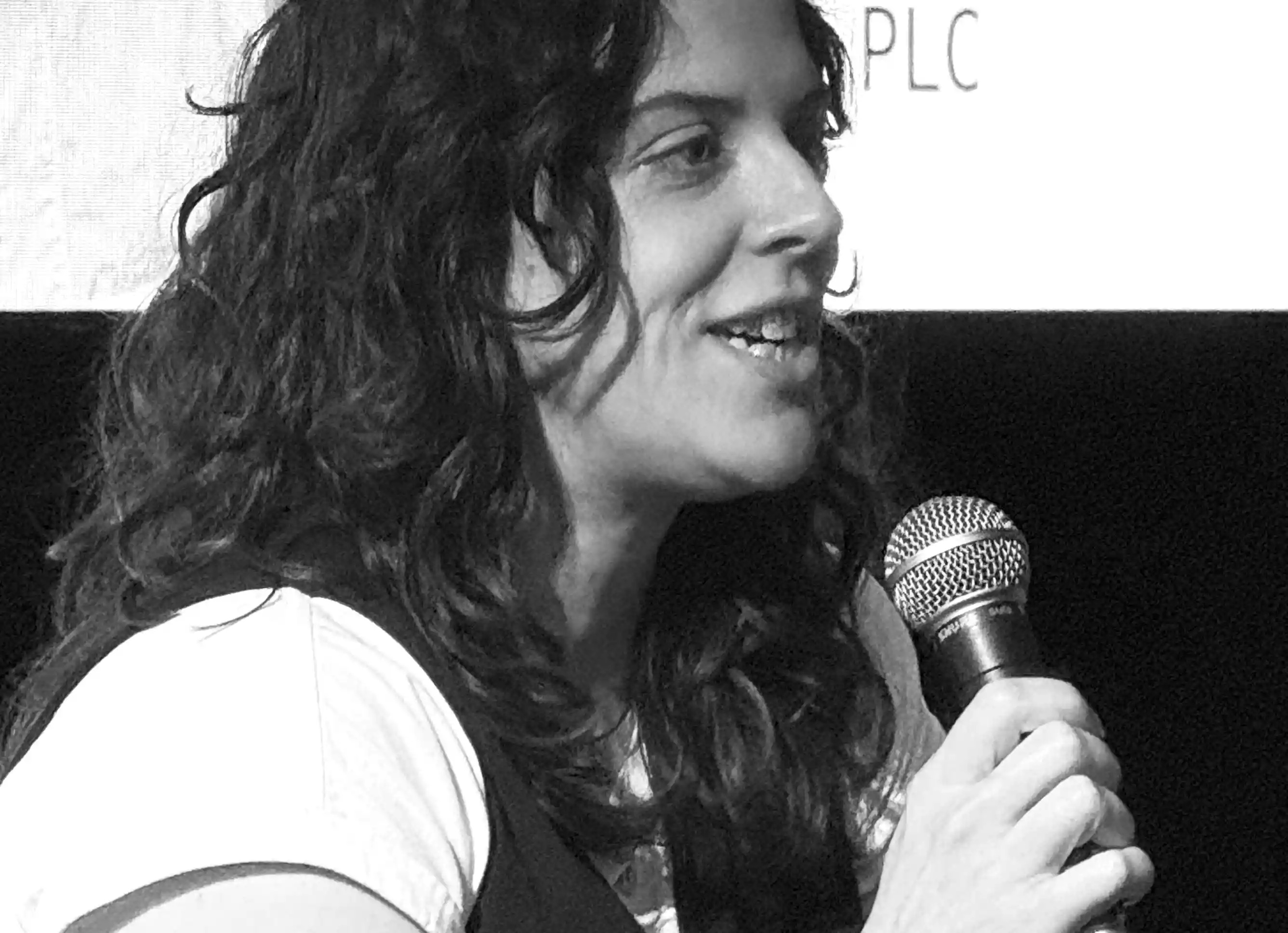 Woman with long curly hair speaking into a microphone in a black and white photo.