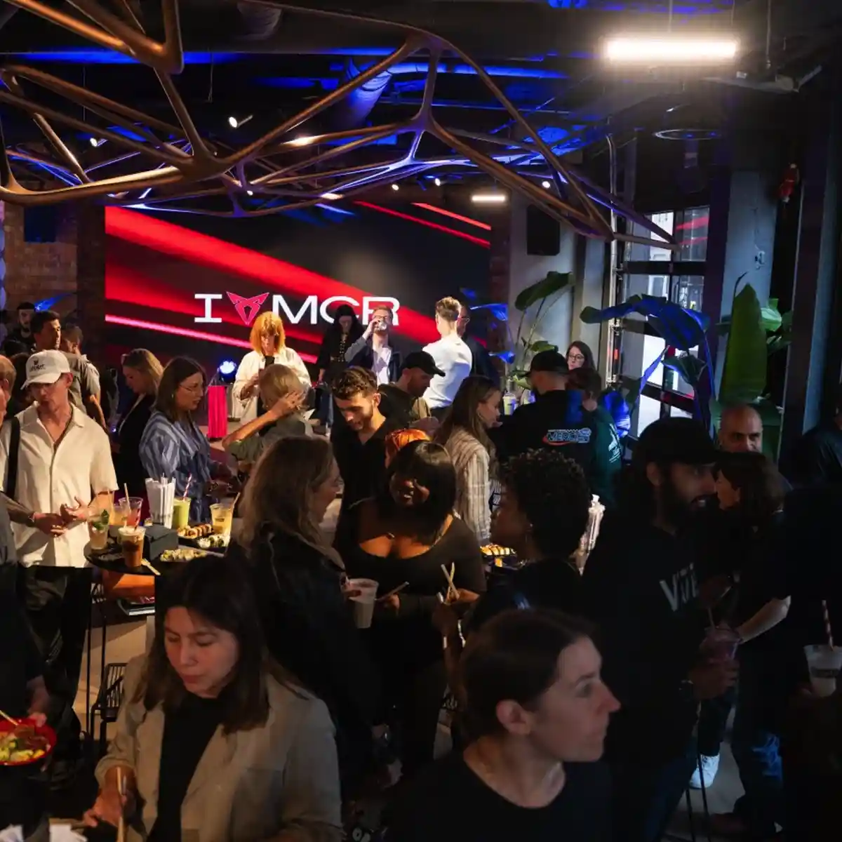 Crowd of people socialising and eating in a modern event space with a large screen showing 'I ♥ MCR'.