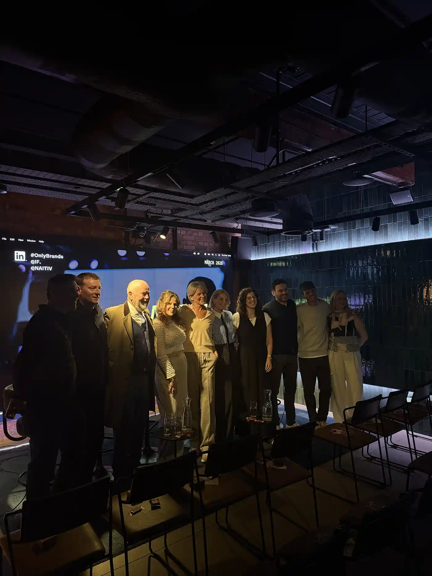 Group of people standing in front of a screen at an indoor event with dim lighting.