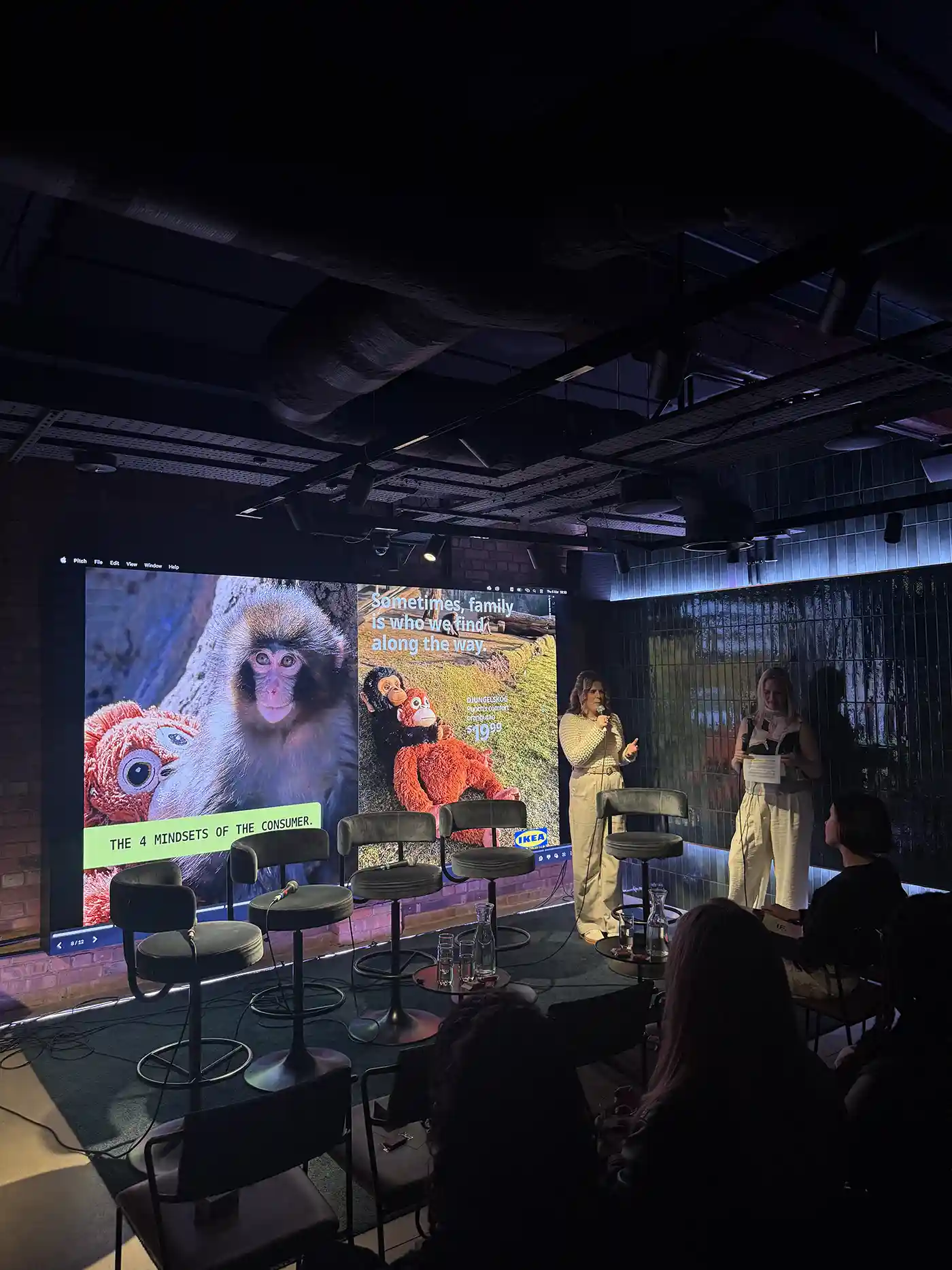 Two speakers standing on a stage next to chairs, with a large screen showing monkey images and text about consumer mindsets.