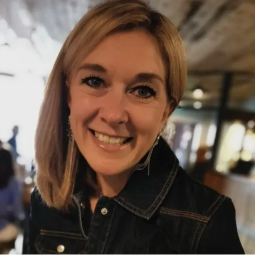 Smiling woman with shoulder-length blonde hair wearing a dark denim jacket indoors.