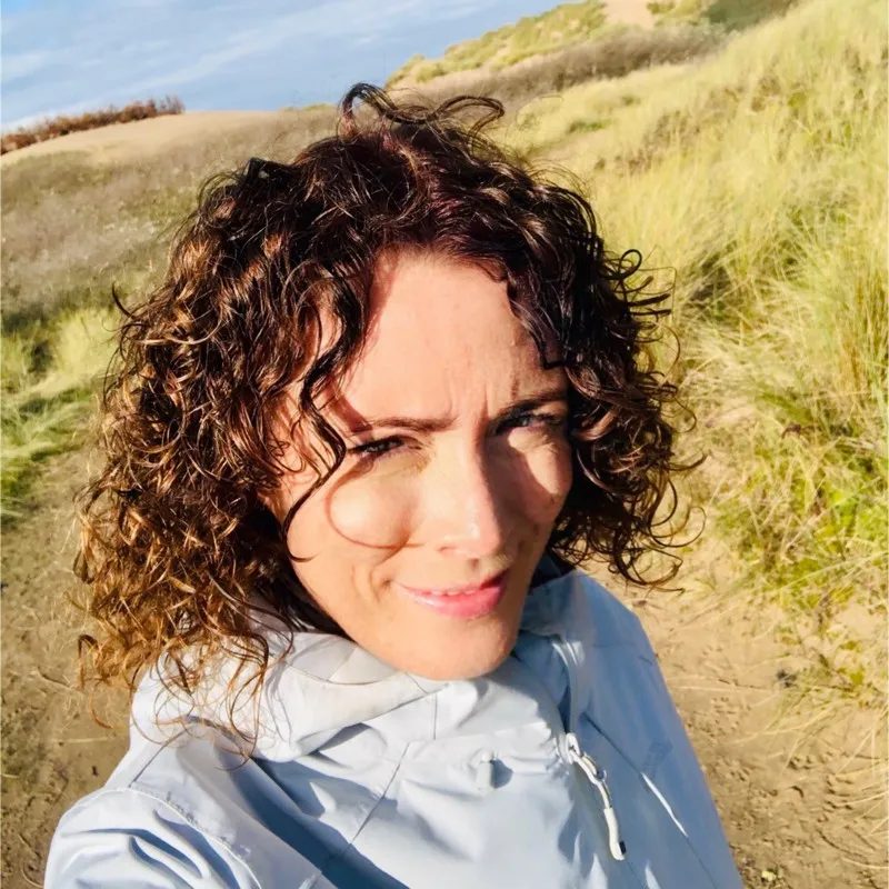 Woman with curly brown hair wearing a light jacket outdoors on a sunny day in a grassy area.