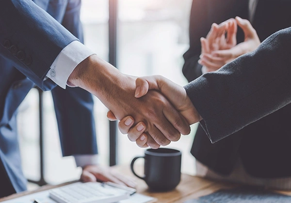 Two people shaking hands across a desk with documents and a laptop.