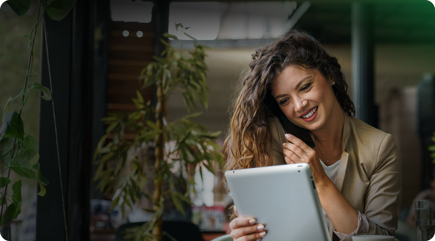 Smiling woman with curly hair in a tan blazer talking on a phone while holding a tablet in a cozy indoor setting with plants.
