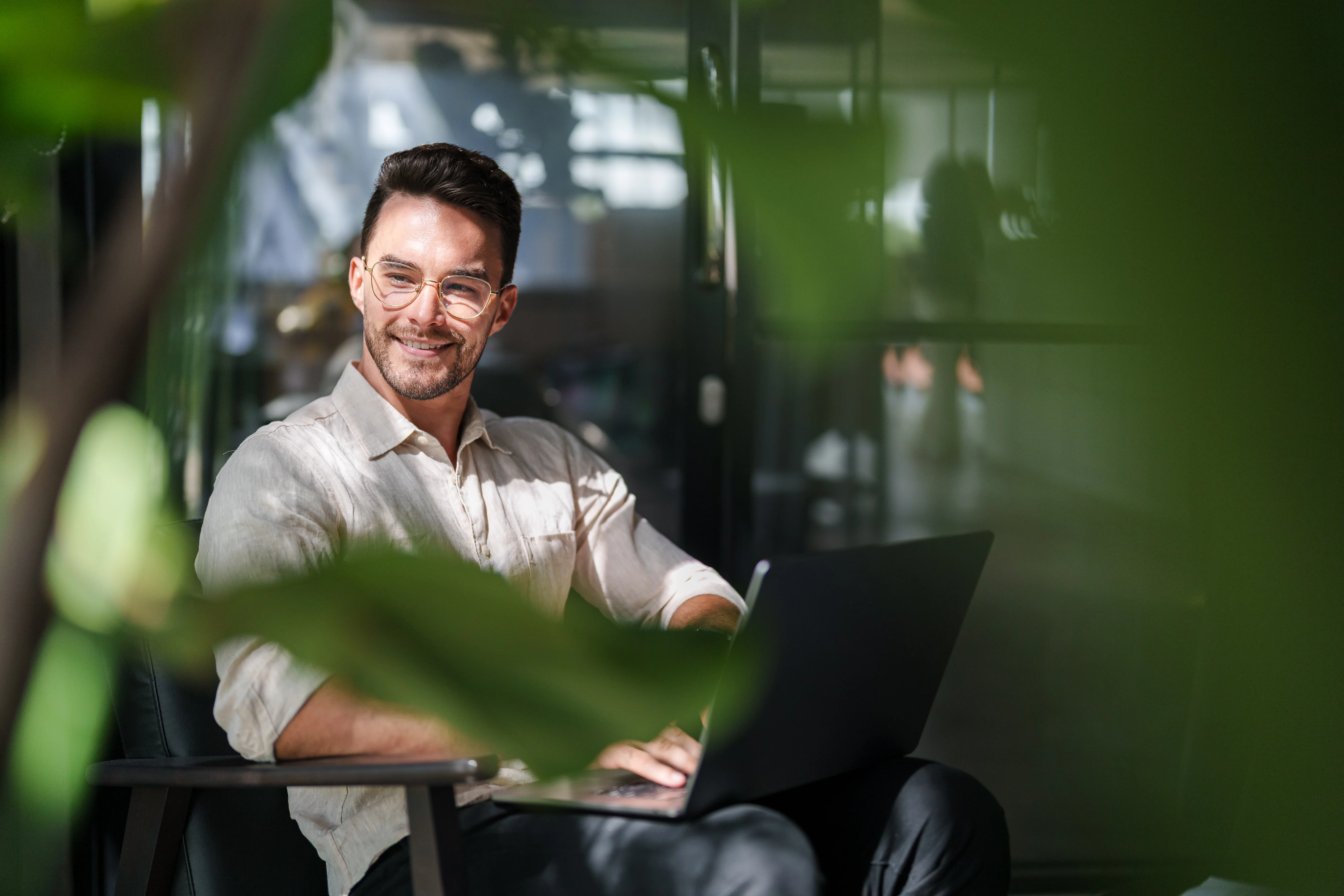 Smiling man wearing glasses sitting indoors with a laptop, partially obscured by green plant leaves.